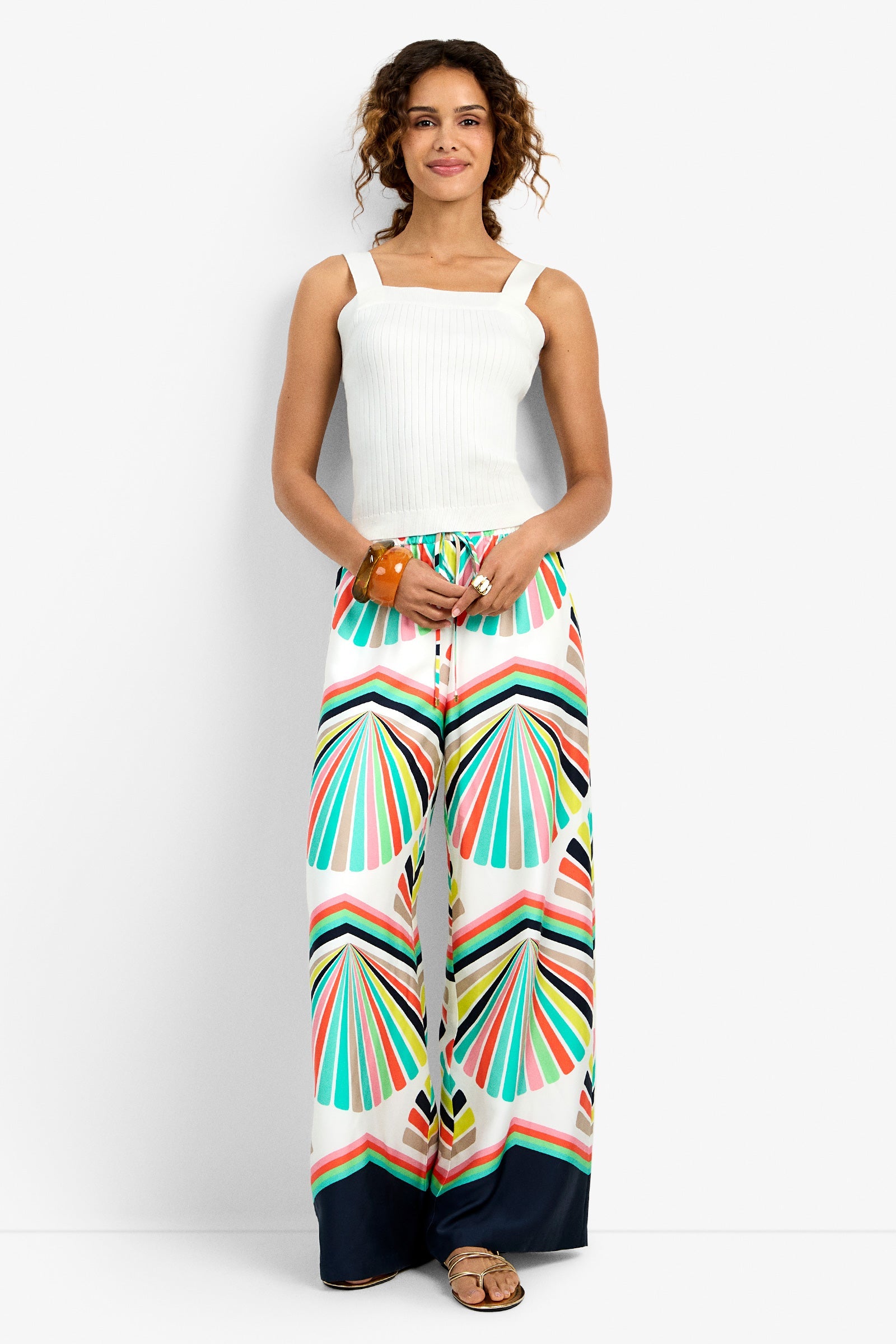Woman standing and smiling, wearing a white ribbed tank top and wide-leg pants with a colorful geometric pattern in teal, coral, and yellow. She has curly hair and is wearing sandals and a wooden bracelet.