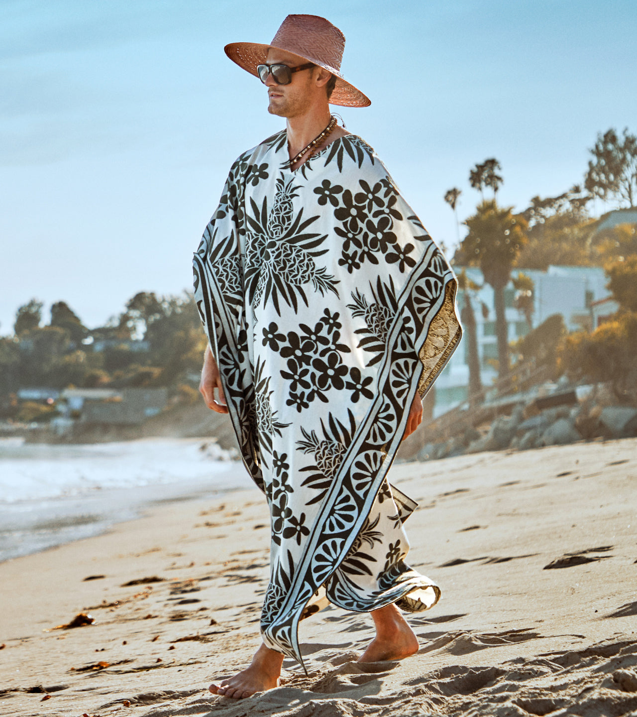A person in a wide-brimmed pink hat and a black-and-white floral kaftan walks barefoot on a sandy beach with trees and buildings in the background.