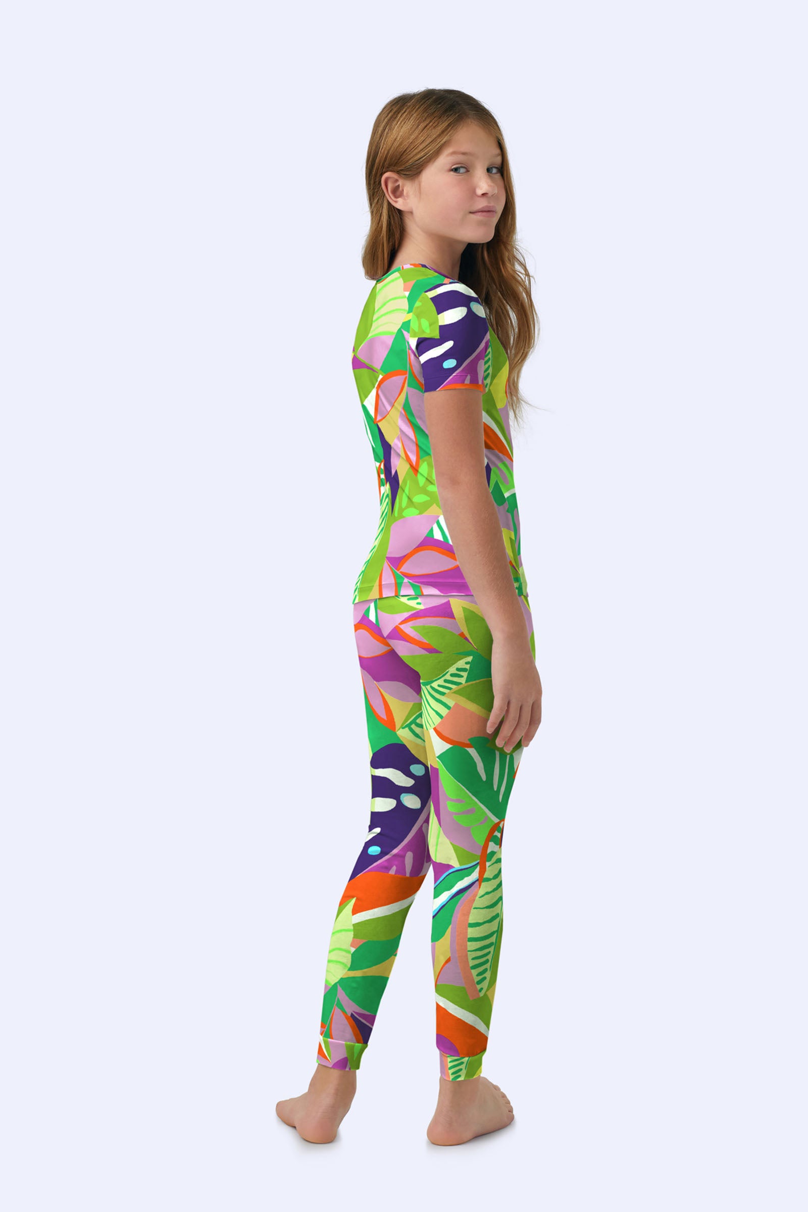 A young girl with long brown hair stands barefoot, slightly turned to the side, wearing the Kids PJ Set - Jungle Stretch Jersey featuring a colorful tropical leaf pattern in green, pink, orange, and purple on a light background.