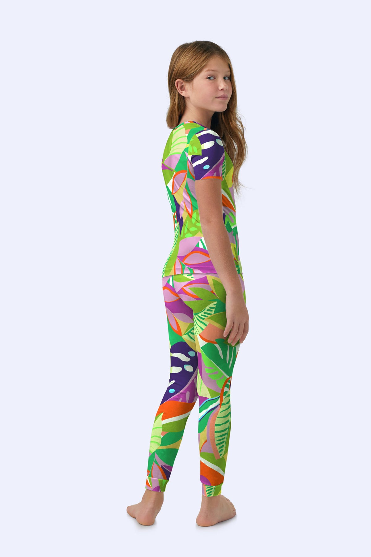 A young girl with long brown hair stands barefoot, slightly turned to the side, wearing the Kids PJ Set - Jungle Stretch Jersey featuring a colorful tropical leaf pattern in green, pink, orange, and purple on a light background.