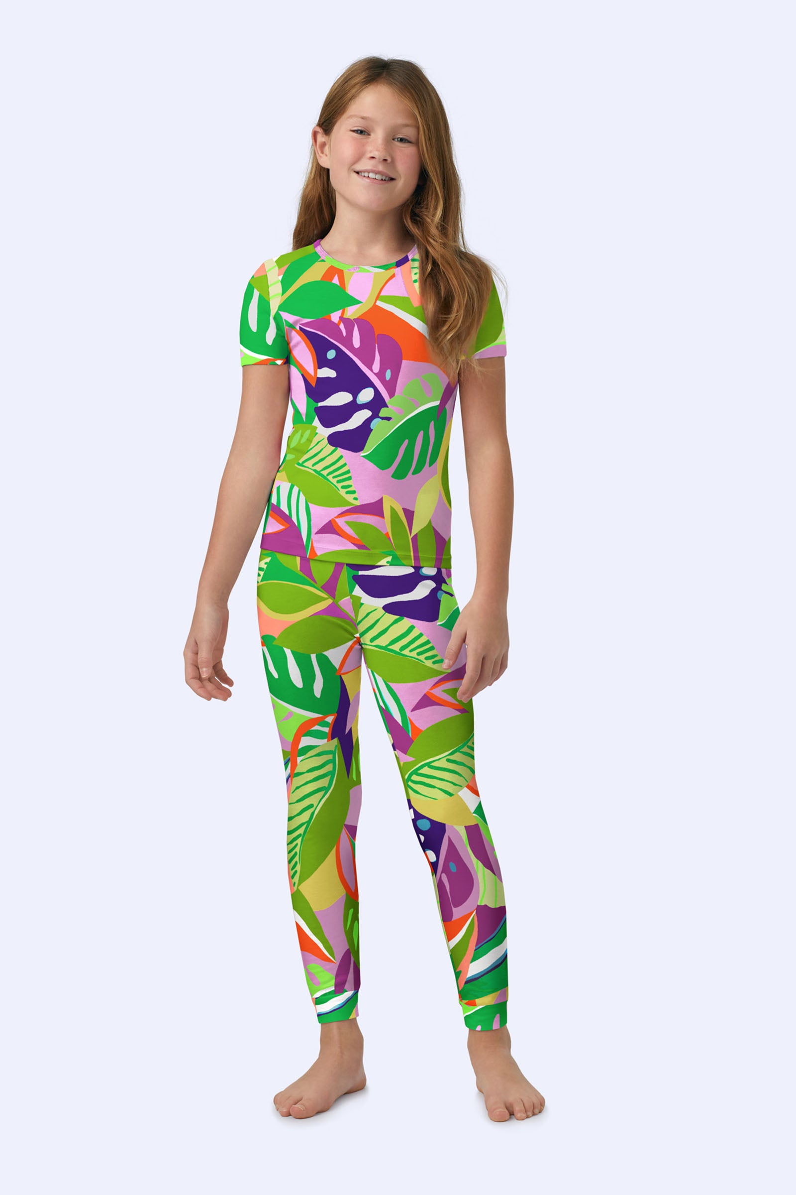 A smiling young girl stands barefoot, wearing the Kids PJ Set - Jungle Stretch Jersey in multi, a colorful fitted outfit with short sleeves and long pants covered in vibrant tropical leaf patterns on a pale background.