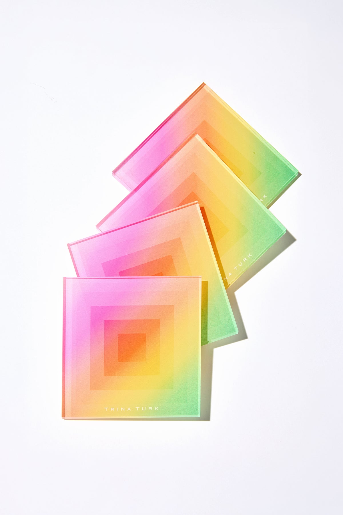 The Acrylic Rainbow Ombre Coaster Set with Holder features four colorful square coasters in a rainbow gradient, stacked diagonally on a white surface. The bottom coaster displays Trina Turk in gold text.