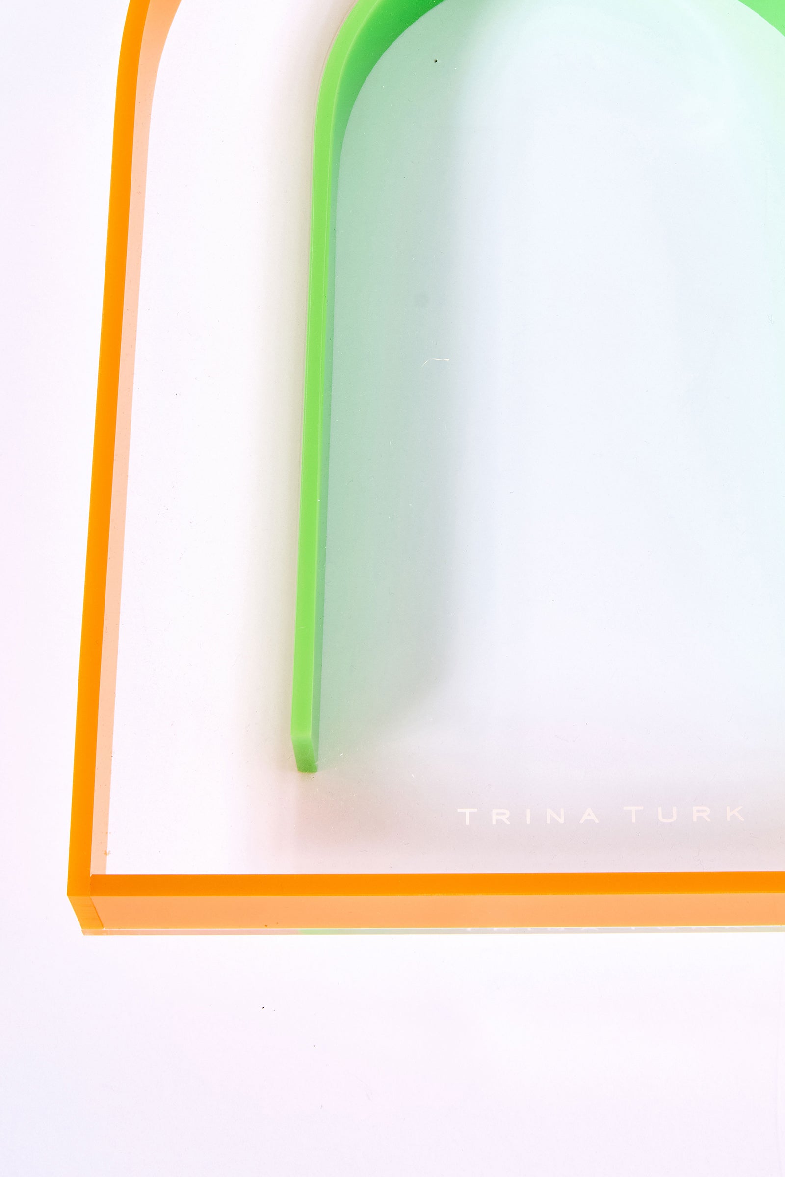 A close-up photo of the Arched Acrylic Serving Tray, a transparent rectangular piece with orange and green curved edges and “Trina Turk” in white text on the bottom right corner.