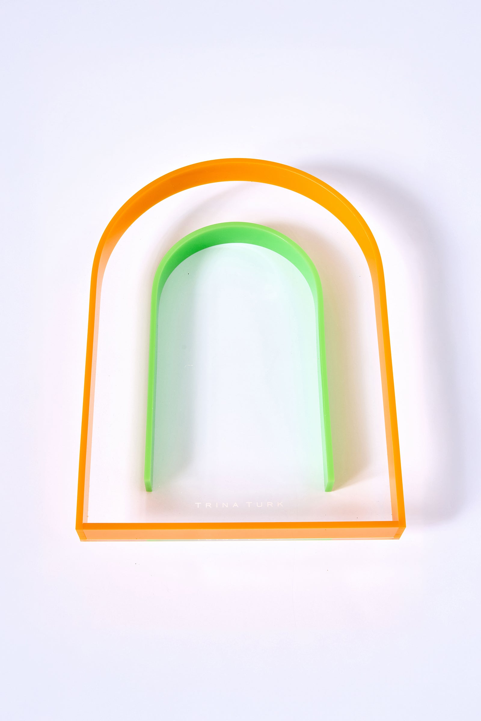 The Arched Acrylic Serving Tray is a clear, rectangular tray with rounded top edges, an orange border, and a green U-shaped curve inside. Trina Turk appears subtly near the bottom center.