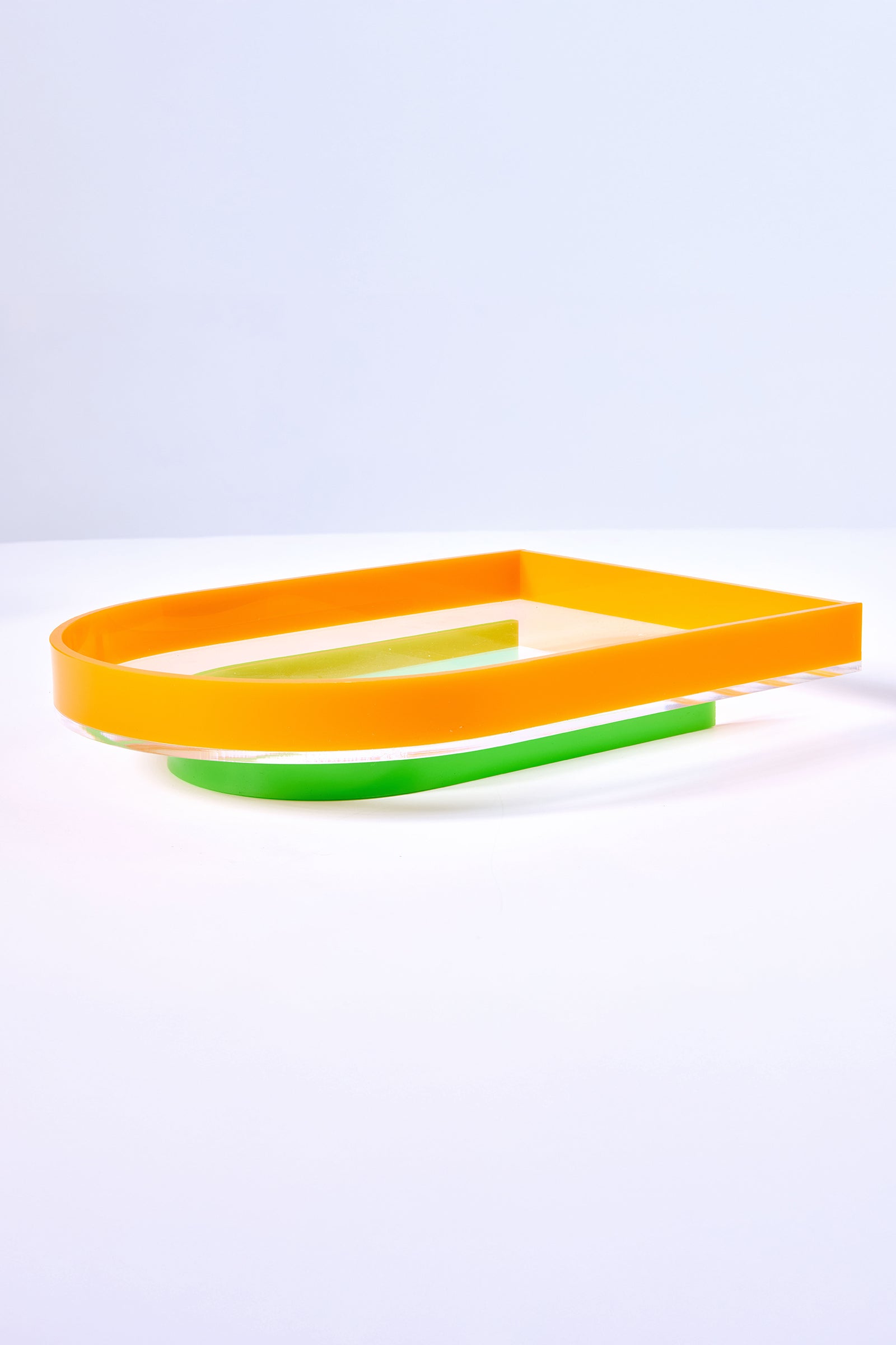 The Arched Acrylic Serving Tray features a minimalist design with a flat, rectangular yellow-orange frame atop a curved green base, all set against a white surface and light blue background.