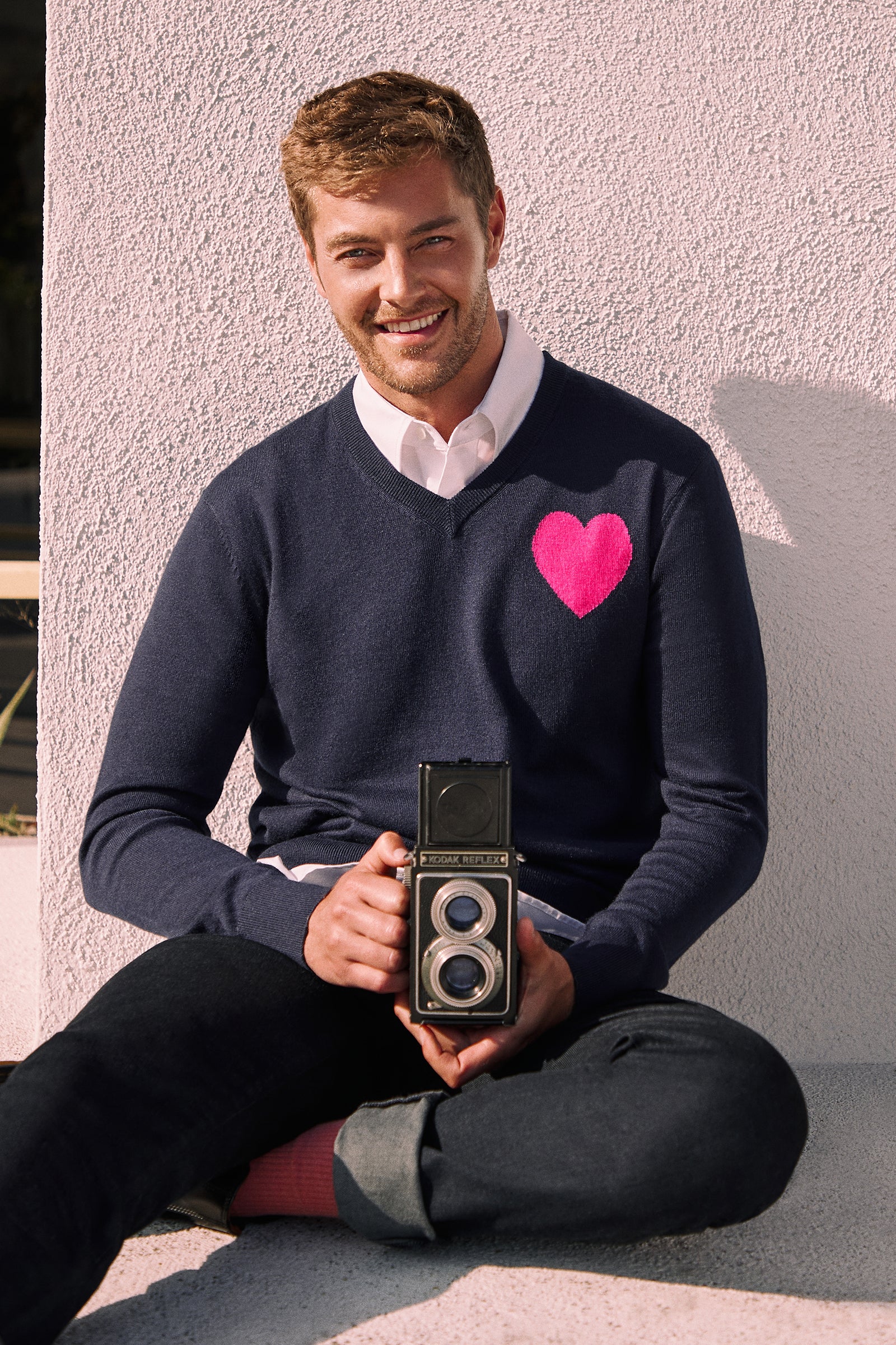 A smiling man sits cross-legged outdoors against a light wall, wearing the Lover Heart V-Neck Sweater in navy with a pink heart on the chest, pink socks, and holding a vintage twin-lens camera in his lap.