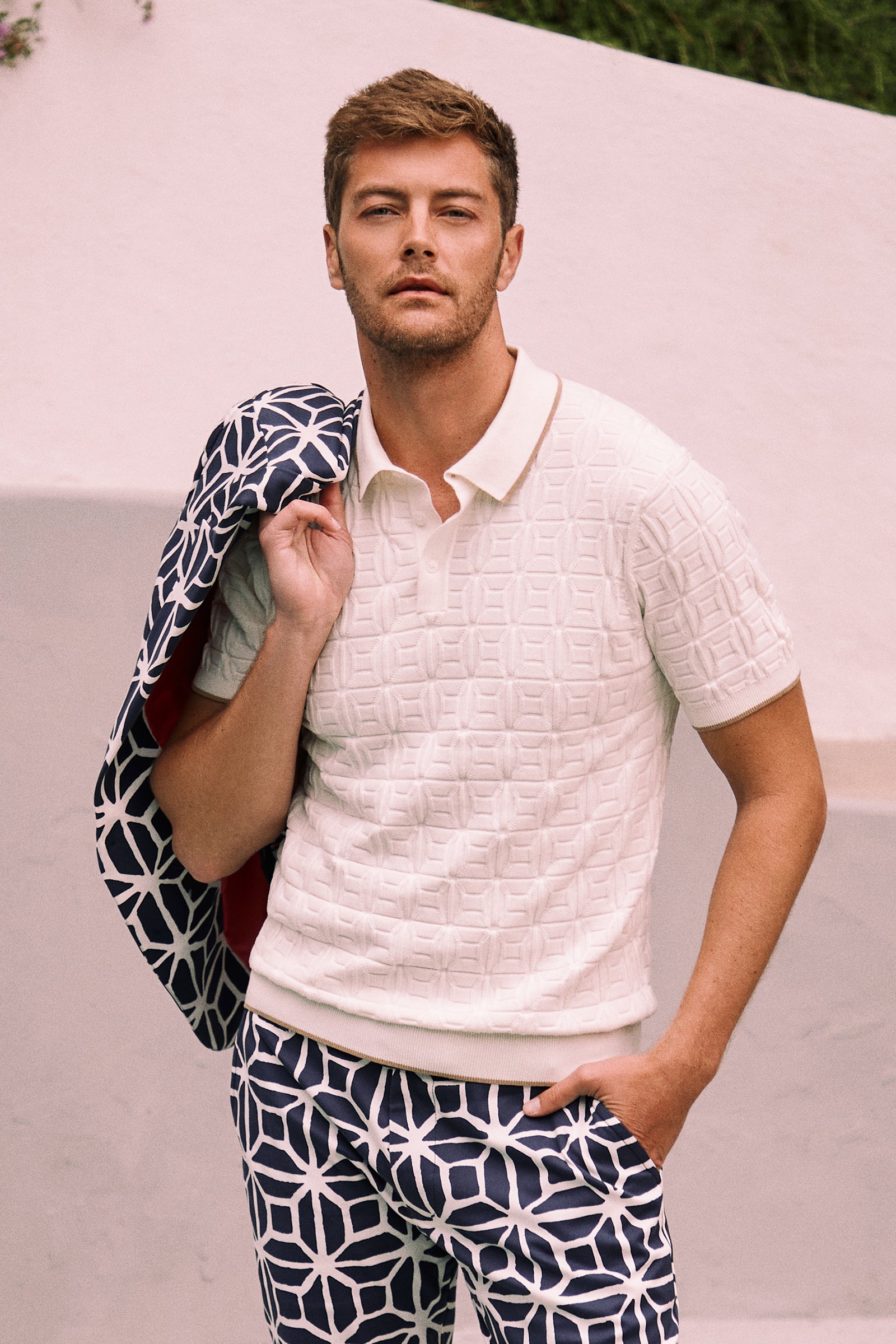 A man poses outdoors with a patterned jacket over his shoulder, wearing the Camilo Short-Sleeve Polo in textured white and navy pants featuring a bold geometric white pattern.