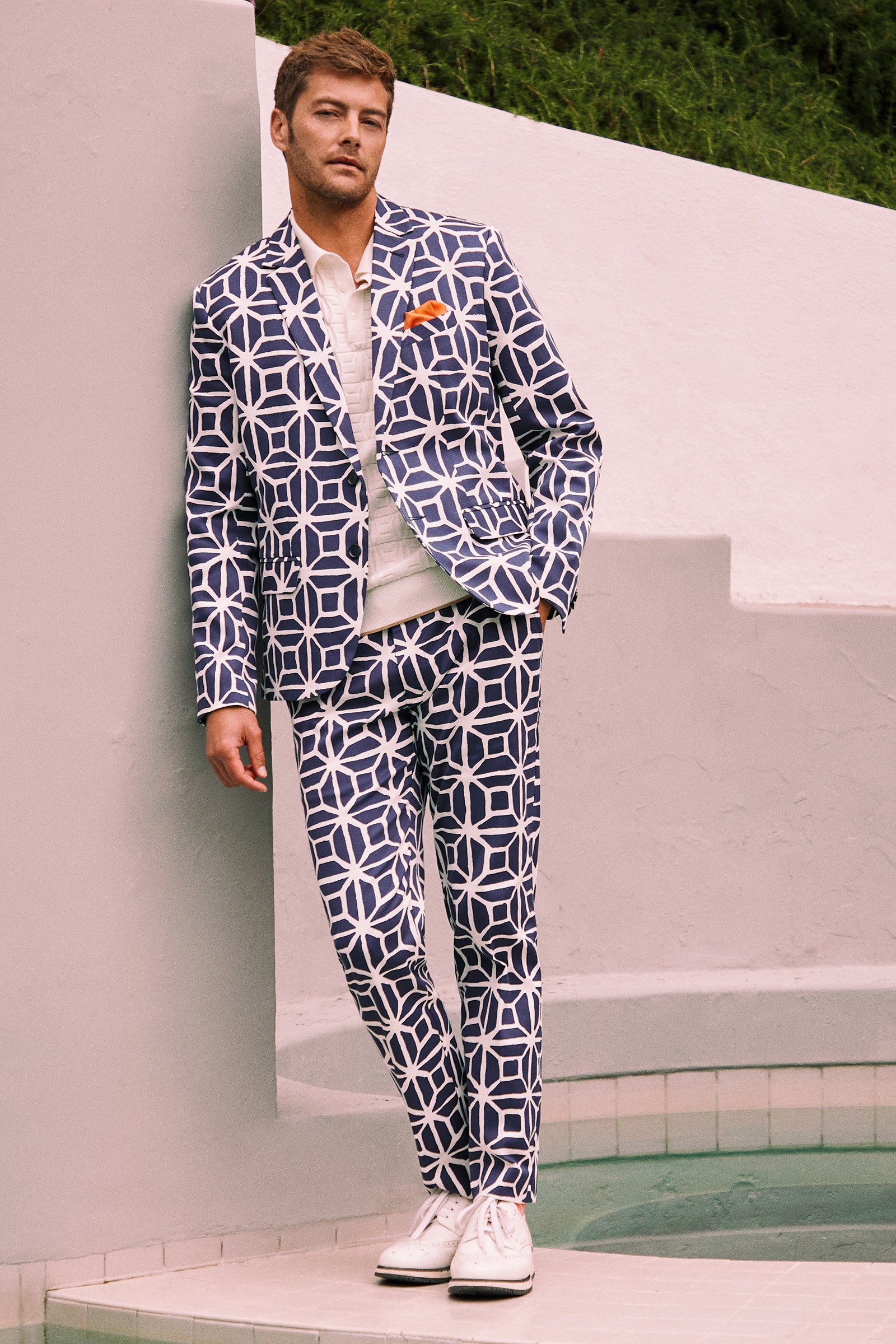 A man stands by a pool in Clyde Slim Trouser - Durell Herringbone, styled with a white shirt, white sneakers, an orange pocket square, against white walls and green grass.
