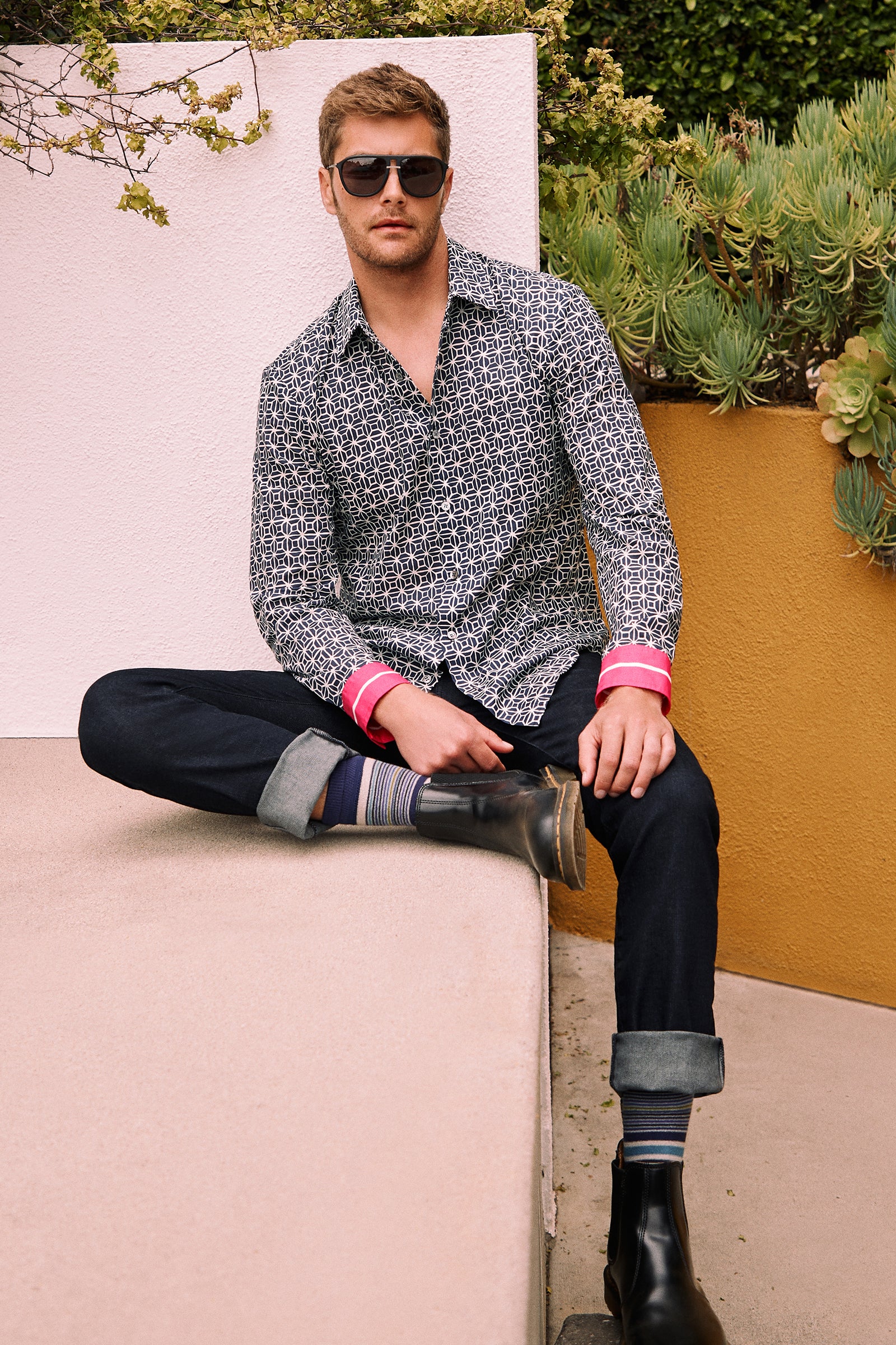 Wearing the Aaron 2 Slim Long-Sleeve Shirt, a man with sunglasses, dark jeans, striped socks, and black boots sits on a beige ledge outdoors against a yellow wall and plants.