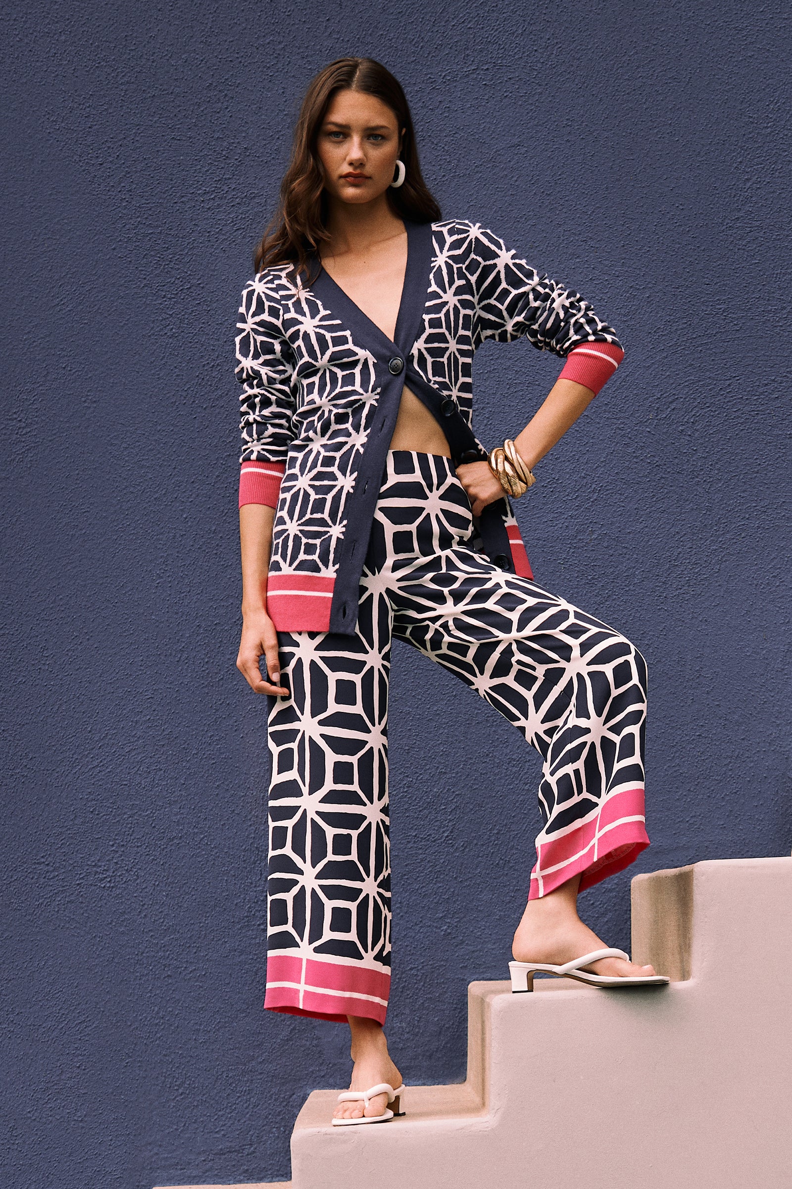 A woman stands on steps by a blue wall, wearing the South Bay V-Neck Cardigan - Durell Knit in a navy and white geometric print, matching wide-leg pants with pink cuffs, white sandals, hoop earrings, and bangles.