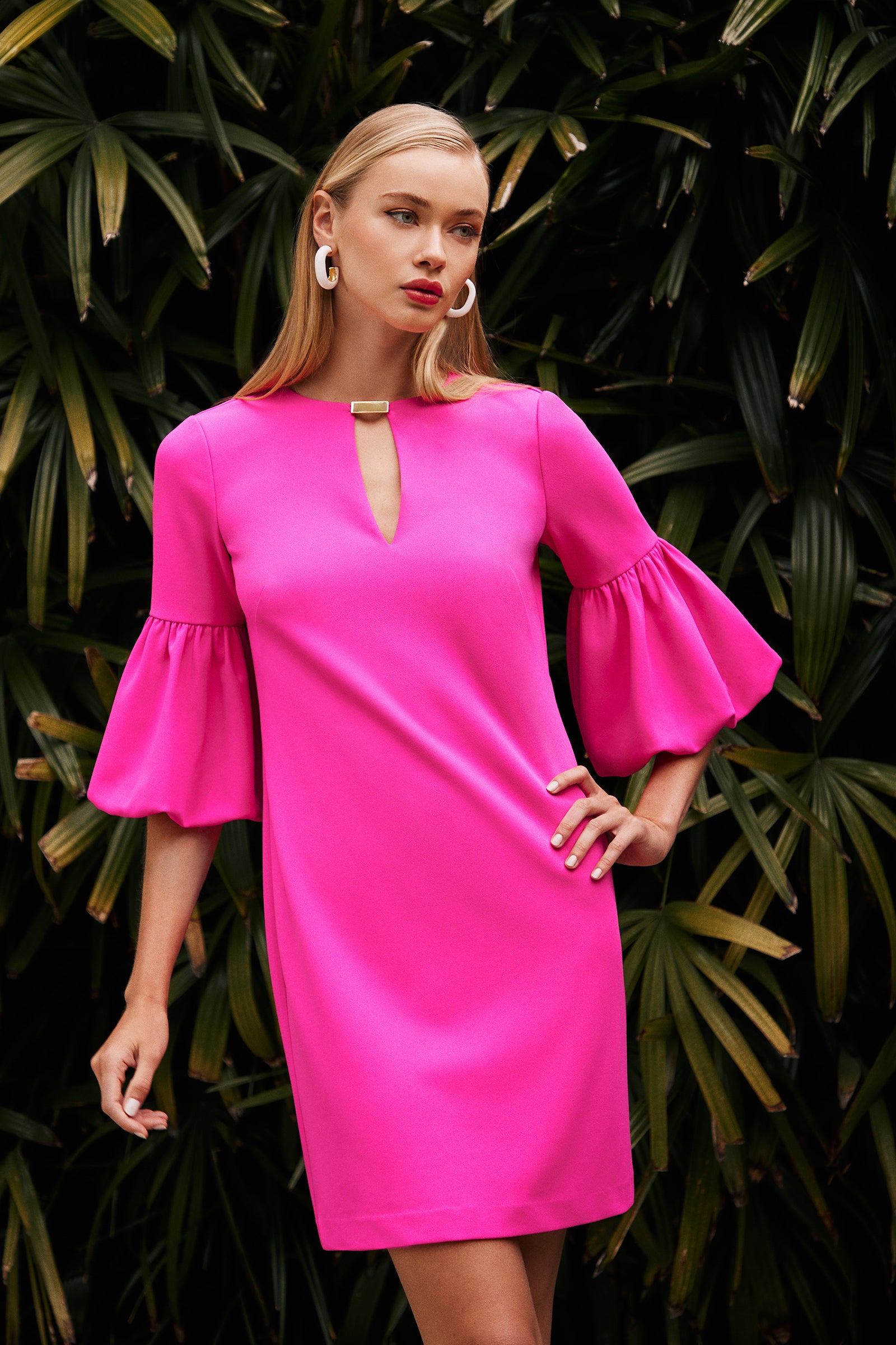 A woman stands confidently among green leafy plants wearing the Redford Dress—a bright pink style with flared sleeves and a front keyhole—paired with bold white earrings.