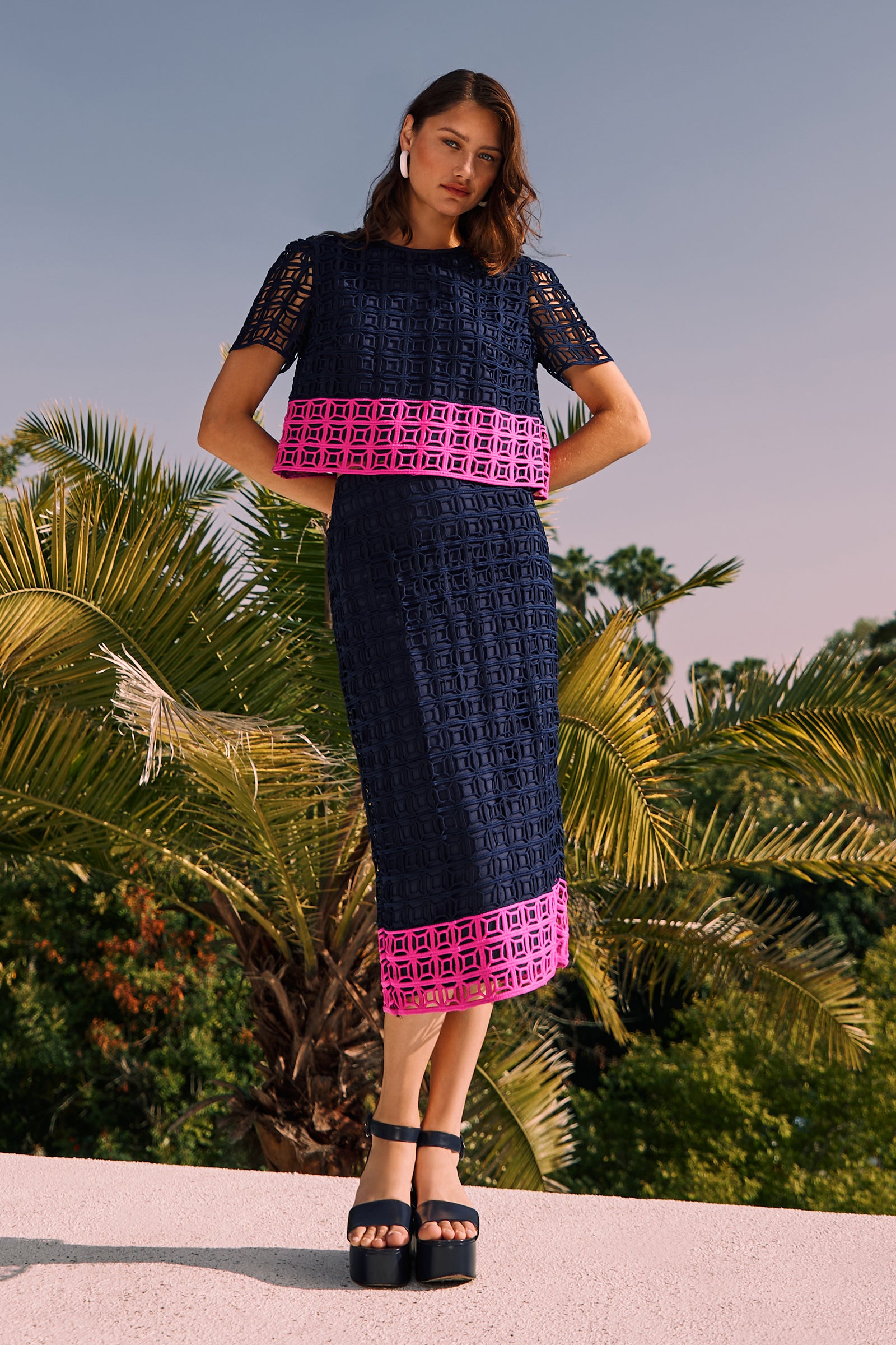 A woman stands outdoors by palm trees, wearing the Dita 2 Midi Pencil Skirt in Block Party Lace—a navy blue geometric patterned skirt with a bright pink hem—paired with black platform sandals.