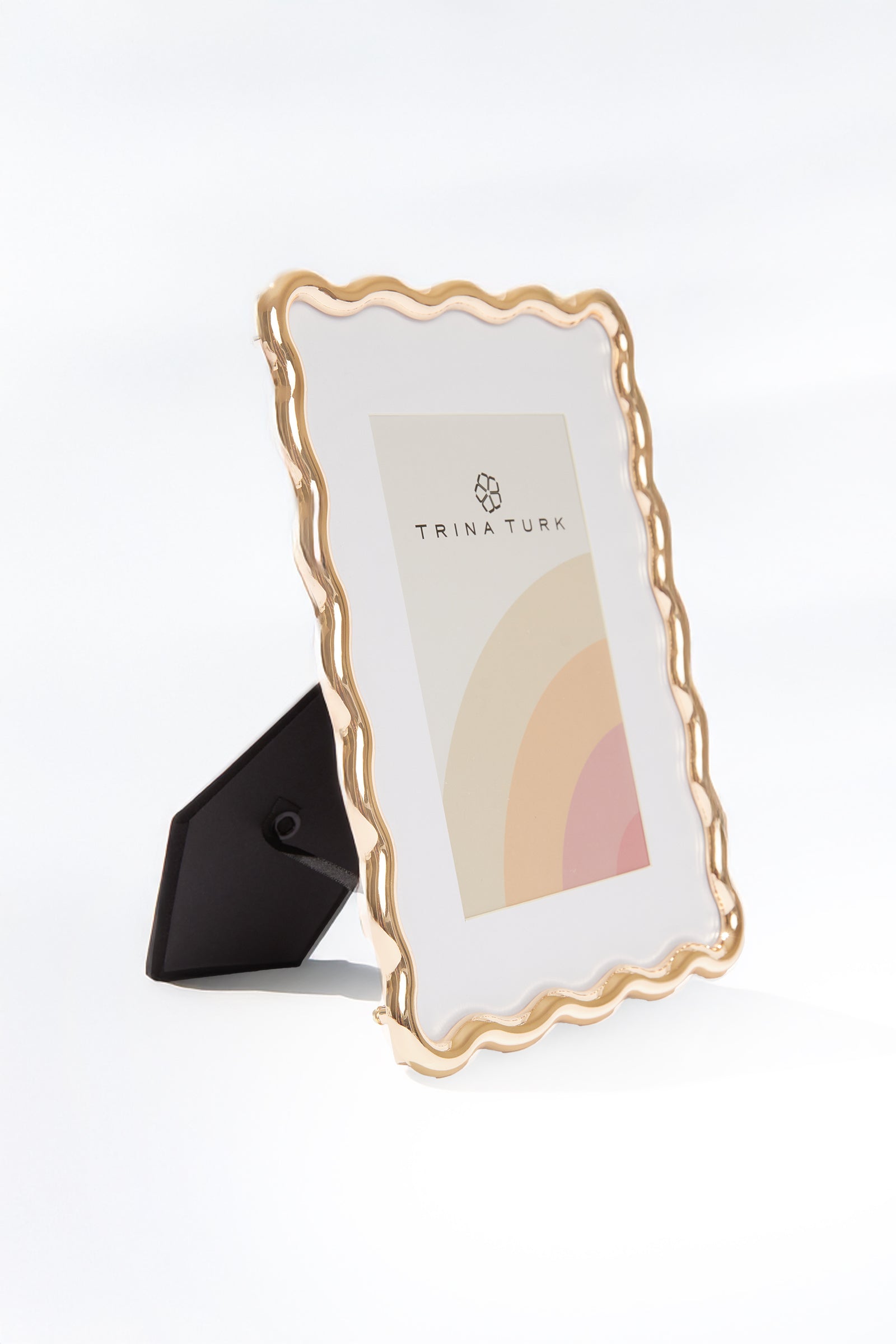 The Trina Turk Wavy Gold Picture Frame features a gold wavy-edged design, fits 4x6 photos, and comes with a stand. It includes a branded placeholder image with a geometric logo and pastel rainbow design on a white background.