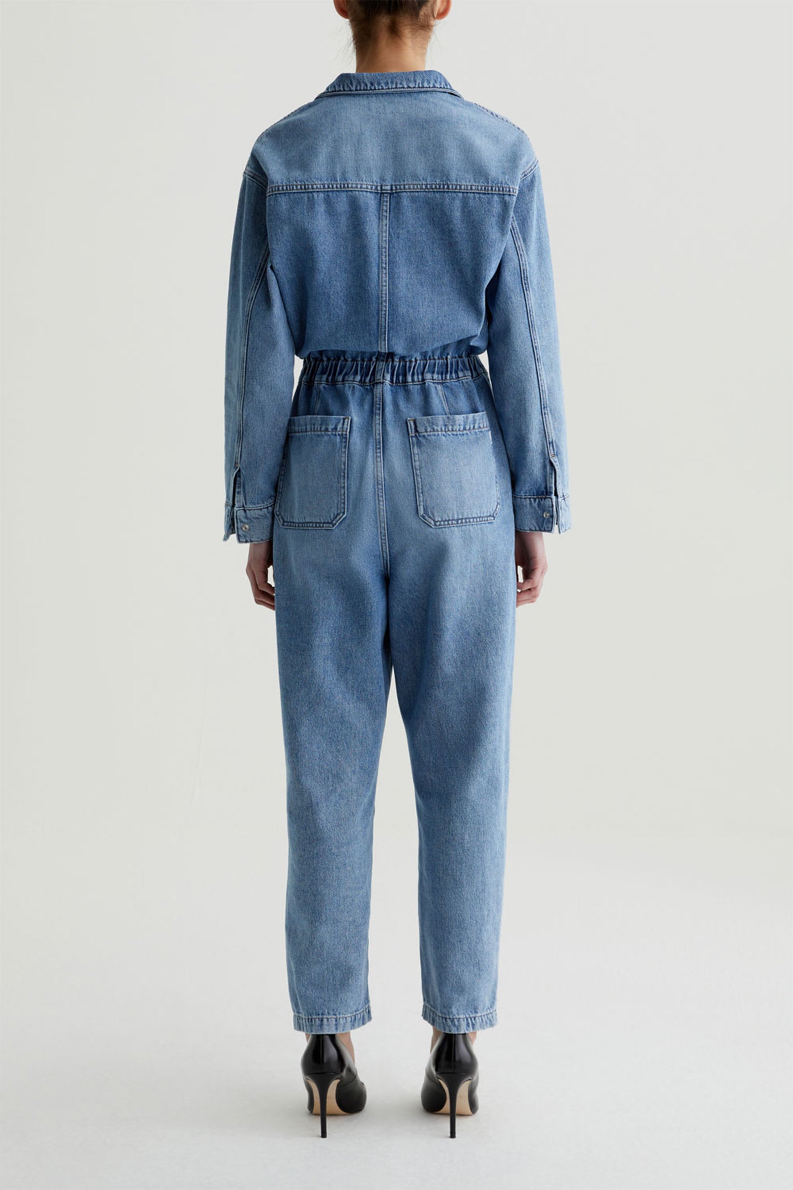 A person stands facing away, wearing the AG Nita Denim Jumpsuit with long sleeves and an elastic waistband, paired with black high heels. The background is plain and light-colored.