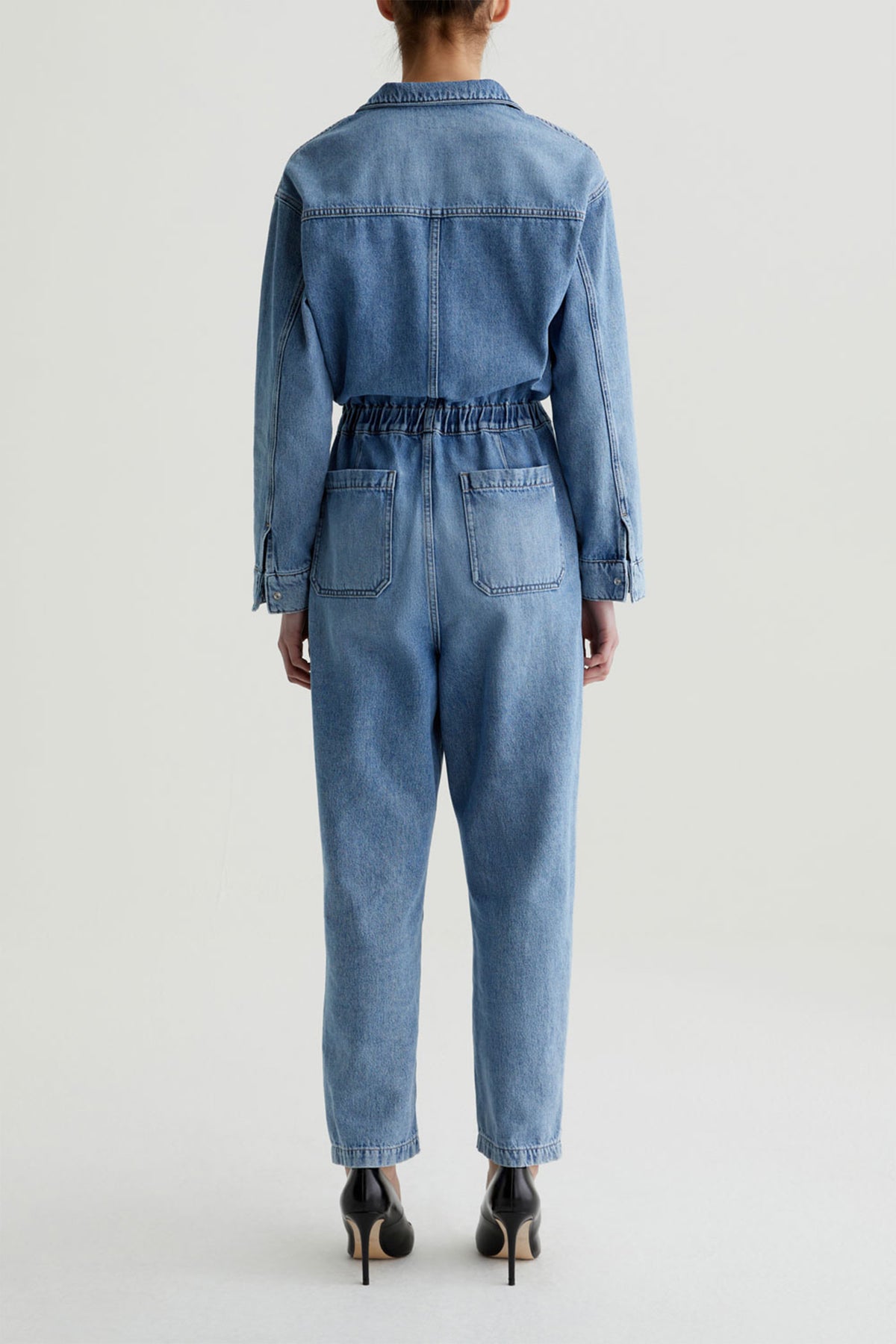 A person stands facing away, wearing the AG Nita Denim Jumpsuit with long sleeves and an elastic waistband, paired with black high heels. The background is plain and light-colored.