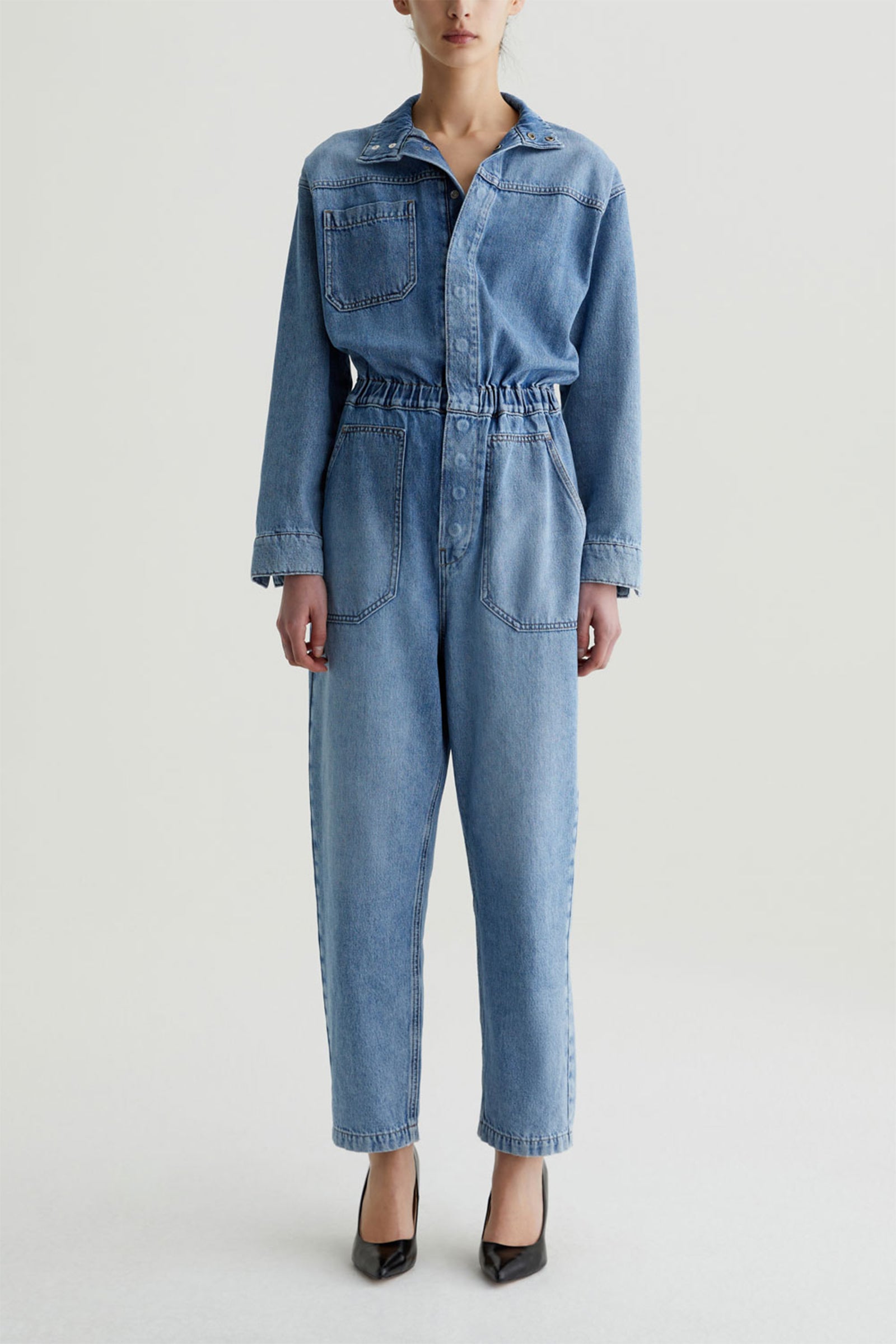 A person stands against a plain background wearing the blue AG Nita Denim Jumpsuit, featuring long sleeves, slouchy denim, and large chest and hip pockets. They pair it with black pointed-toe heels; their face is partially out of frame.