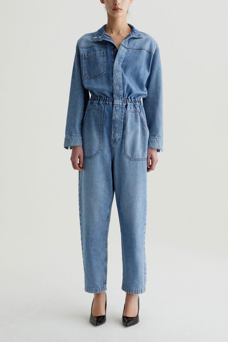 A person stands against a plain background wearing the blue AG Nita Denim Jumpsuit, featuring long sleeves, slouchy denim, and large chest and hip pockets. They pair it with black pointed-toe heels; their face is partially out of frame.