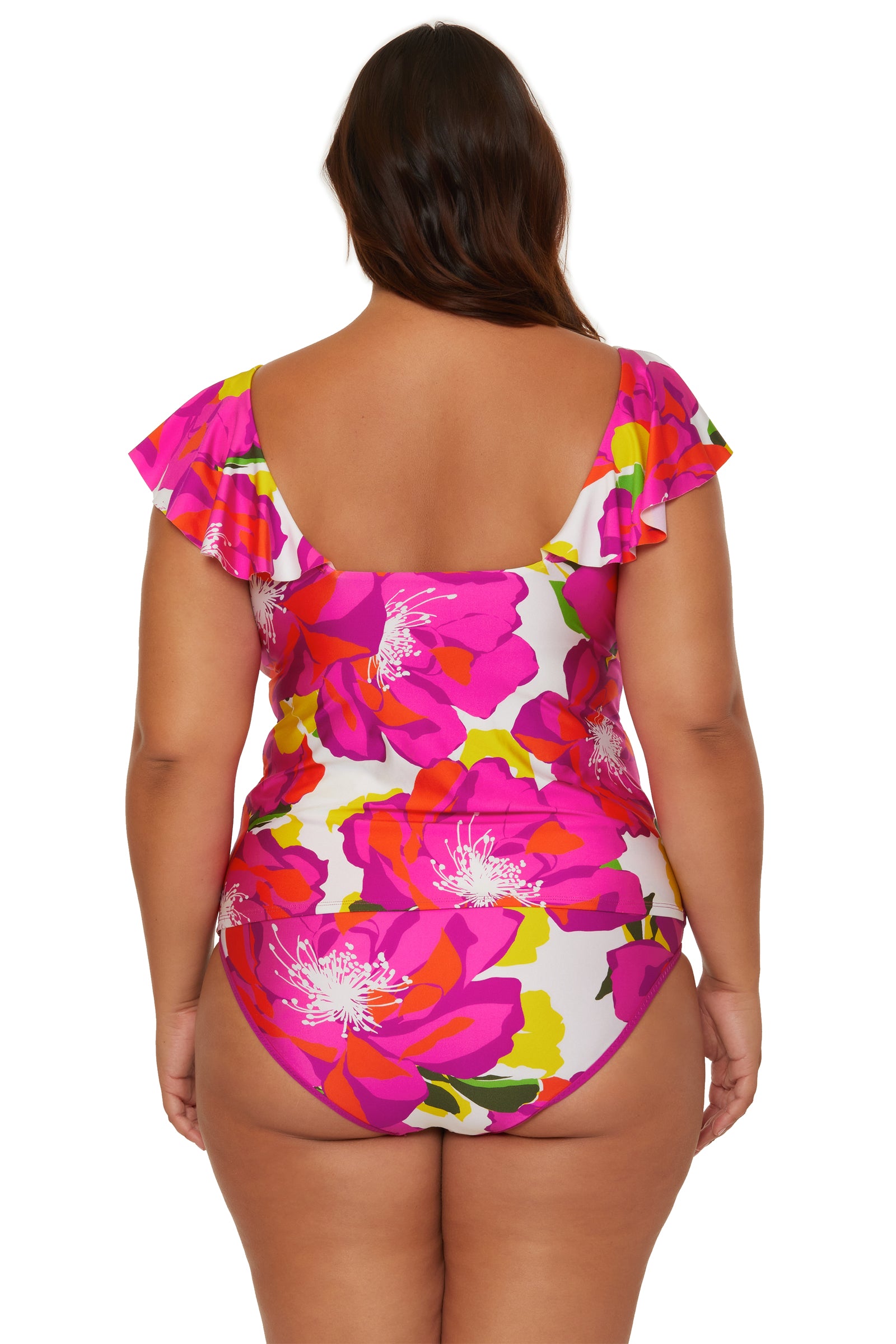 A woman with long brown hair is seen from the back, wearing the Bloom Ruffle Plunge Tankini by Trina Turk in vibrant pink, red, orange, green, and white floral patterns with ruffled short sleeves.