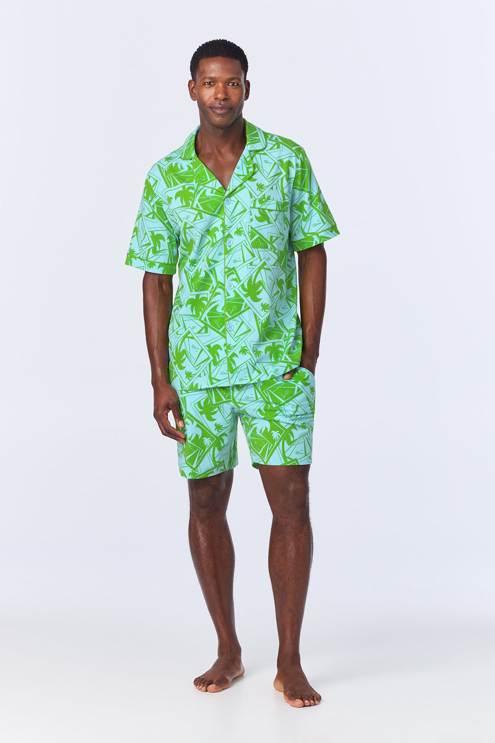 A man stands barefoot against a plain light background wearing the Mens Boxer PJ Set - Tennis Stretch Jersey in blue, featuring a matching short-sleeve button-up shirt and shorts.