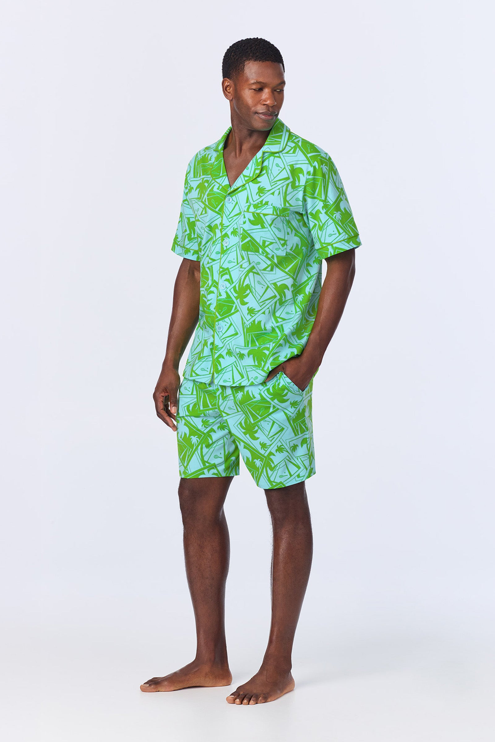 A man stands barefoot with one hand in his pocket, wearing the Mens Boxer PJ Set - Tennis Stretch Jersey, featuring a matching green and blue patterned short-sleeve shirt and shorts, against a plain light background.