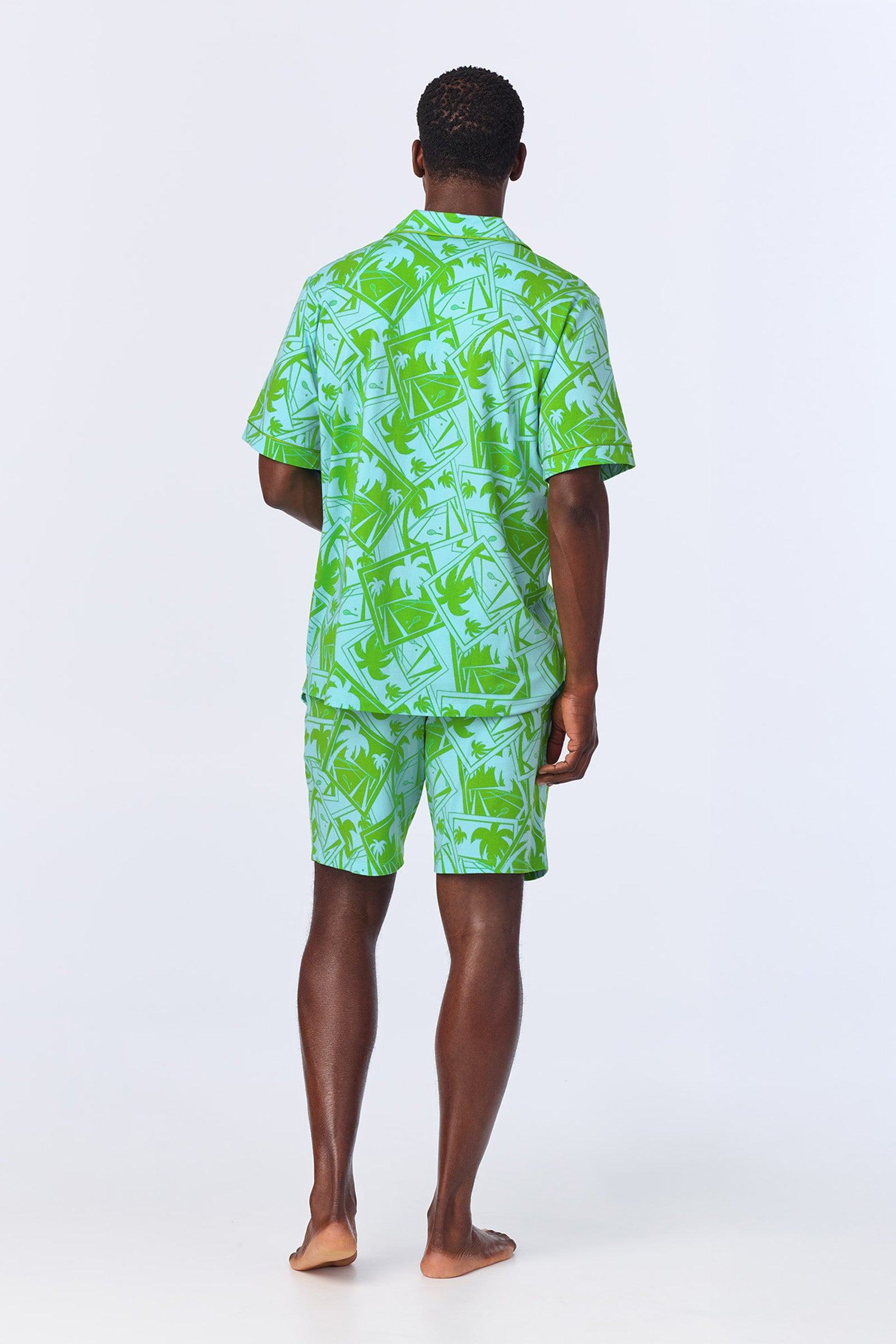 A man stands barefoot facing away, wearing the Mens Boxer PJ Set - Tennis Stretch Jersey featuring a matching green and light blue short-sleeved shirt and shorts with a tropical palm tree and postcard design on a plain background.