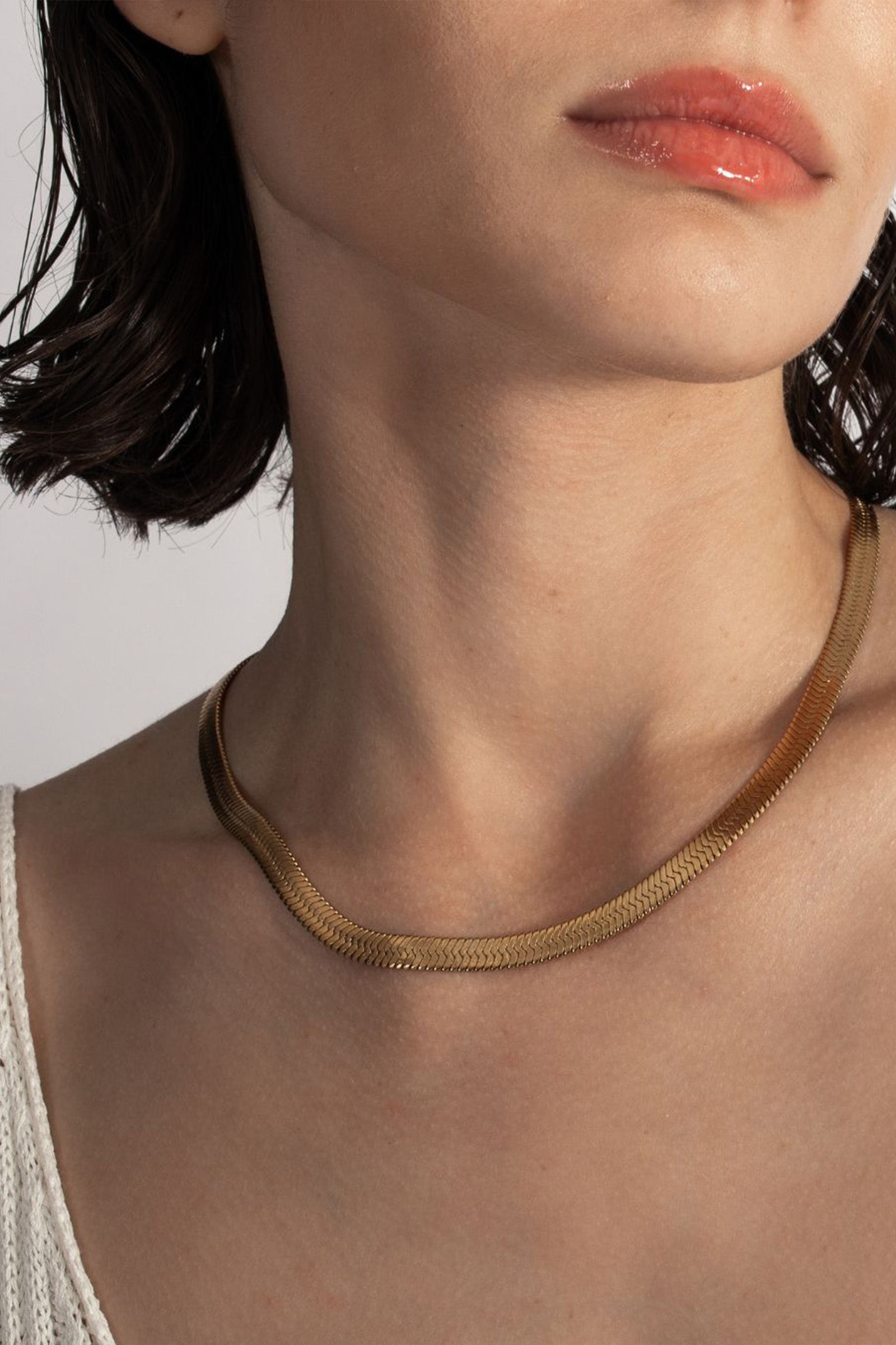 A close-up of a woman with short brown hair and glossy lips, wearing the Celine Lariat gold necklace and a white textured top.
