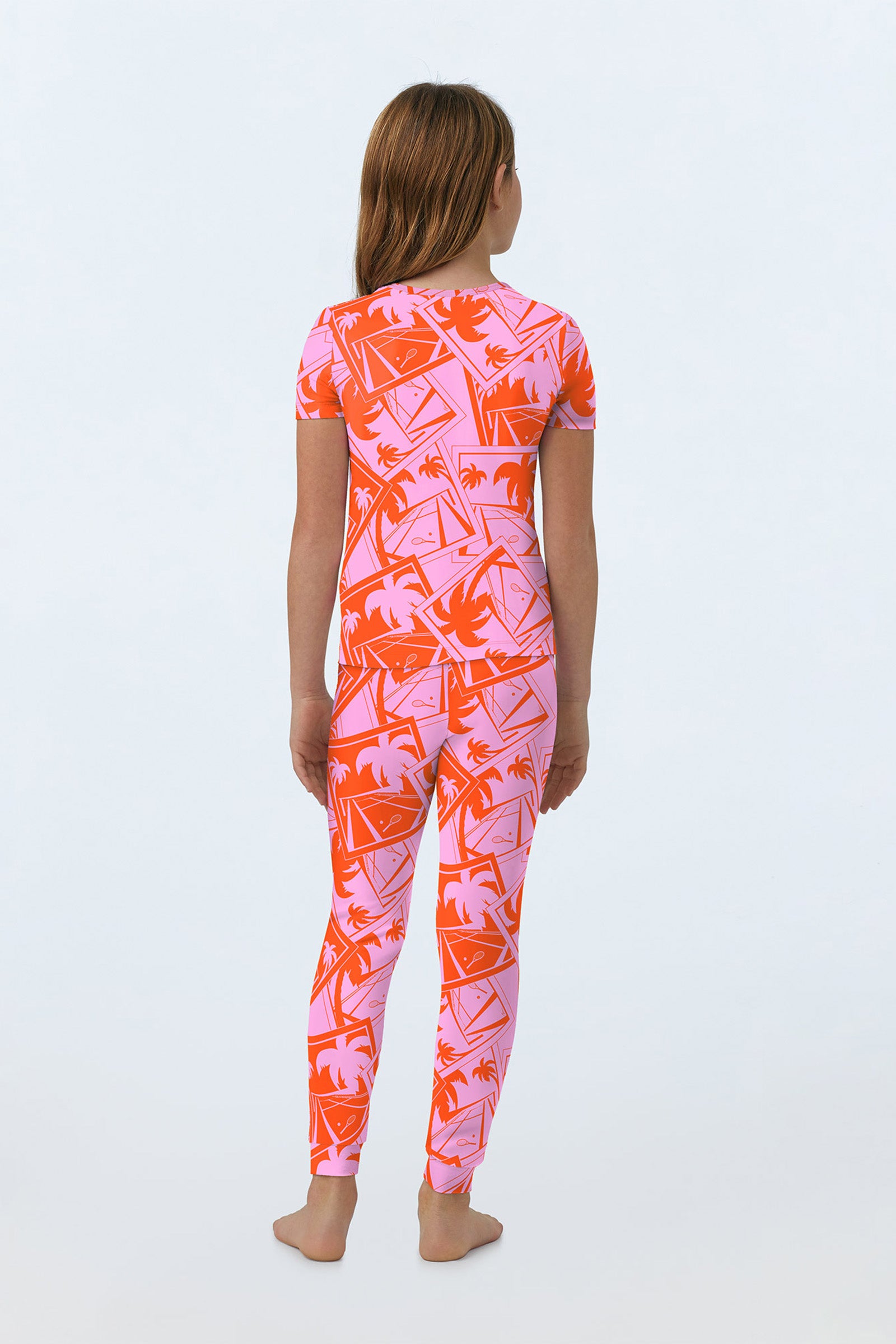 A child stands barefoot facing away, wearing the Pink Kids PJ Set - Tennis Stretch Jersey, a fitted short-sleeved outfit with pink and orange tropical patterns. The background is plain and light-colored.