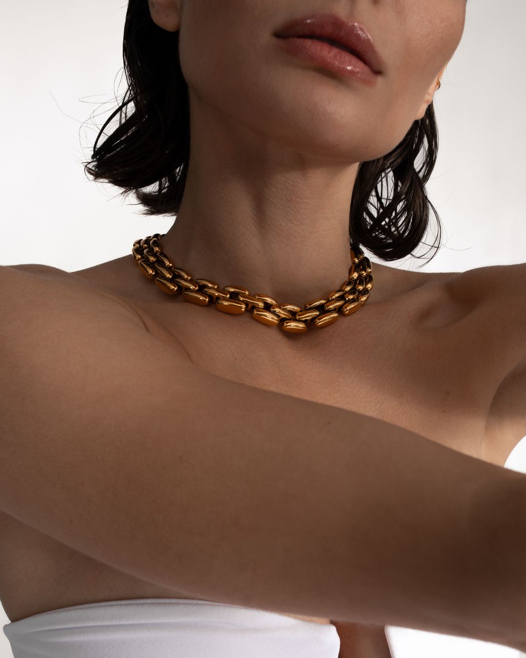 A woman with loose waves in her dark hair poses against a plain background, wearing a white strapless top and the Phoebe 17 Necklace, a chunky gold chain.