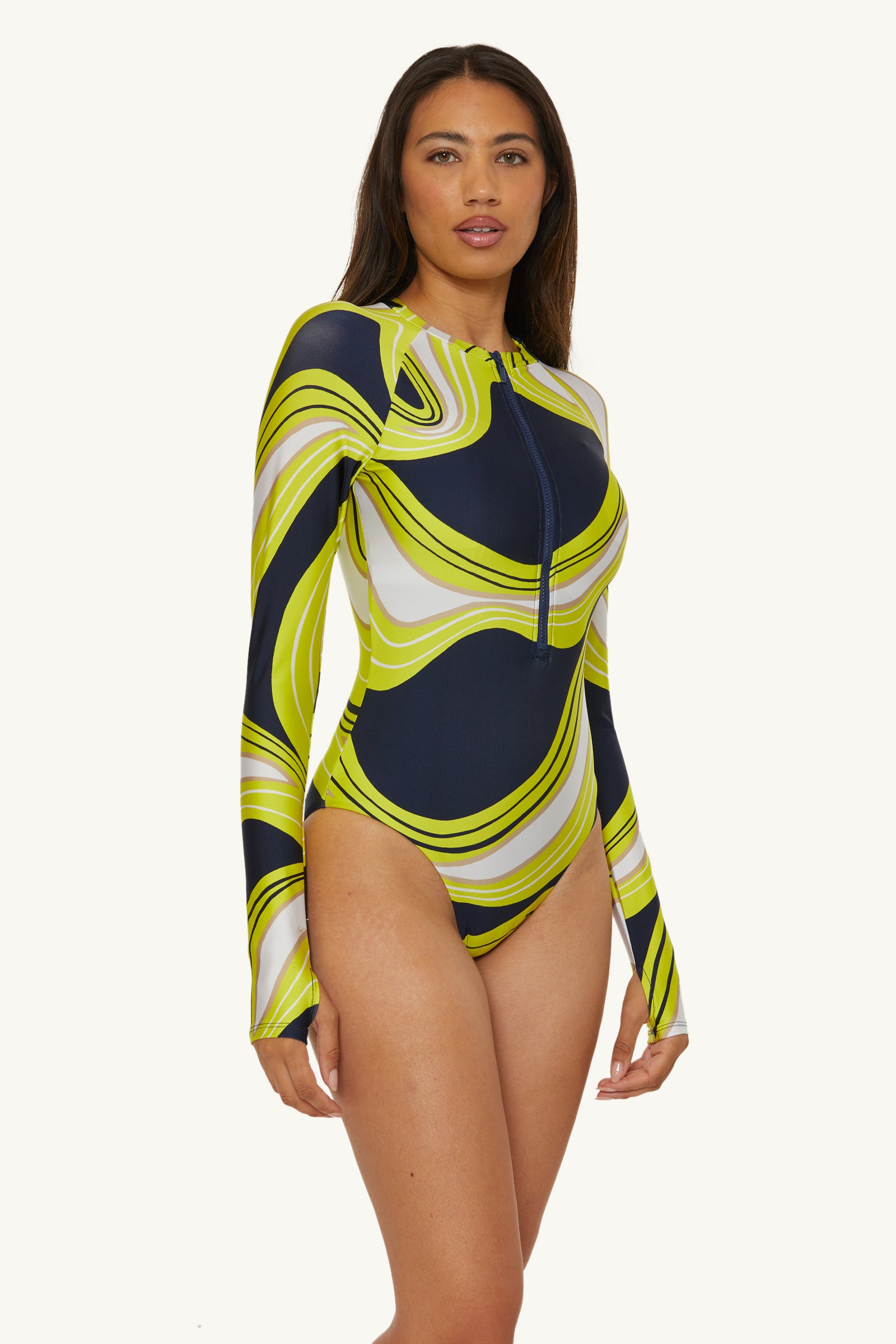 A woman models the Long-Sleeve Zip-Up Paddlesuit - Serena Swirl, featuring a bold wavy pattern in navy, lime green, white, and yellow against a plain light background.