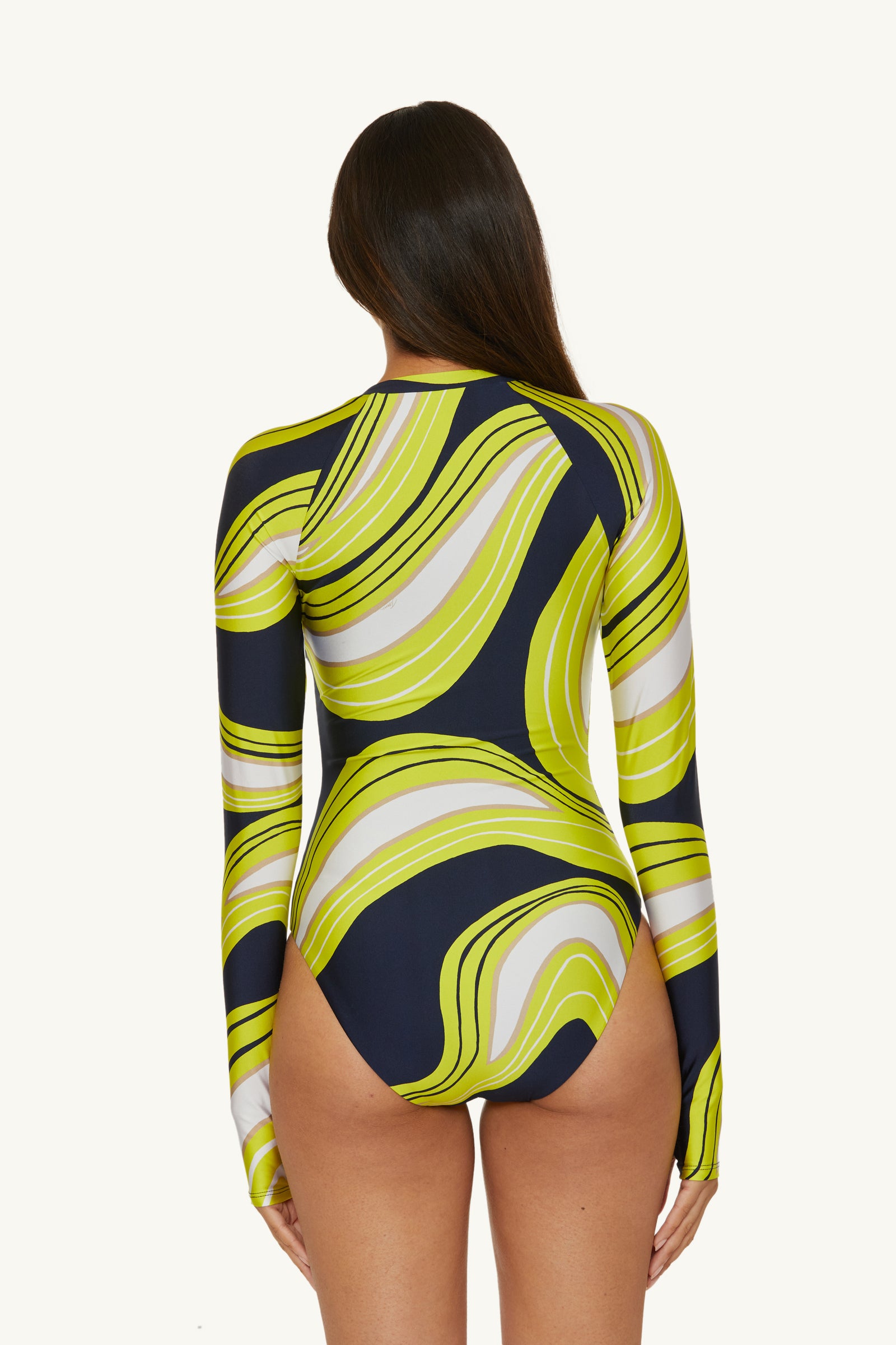 A woman with long brown hair is seen from behind wearing the Serena Swirl Long-Sleeve Zip-Up Paddlesuit, featuring a bold wavy pattern in yellow, green, white, and navy blue.