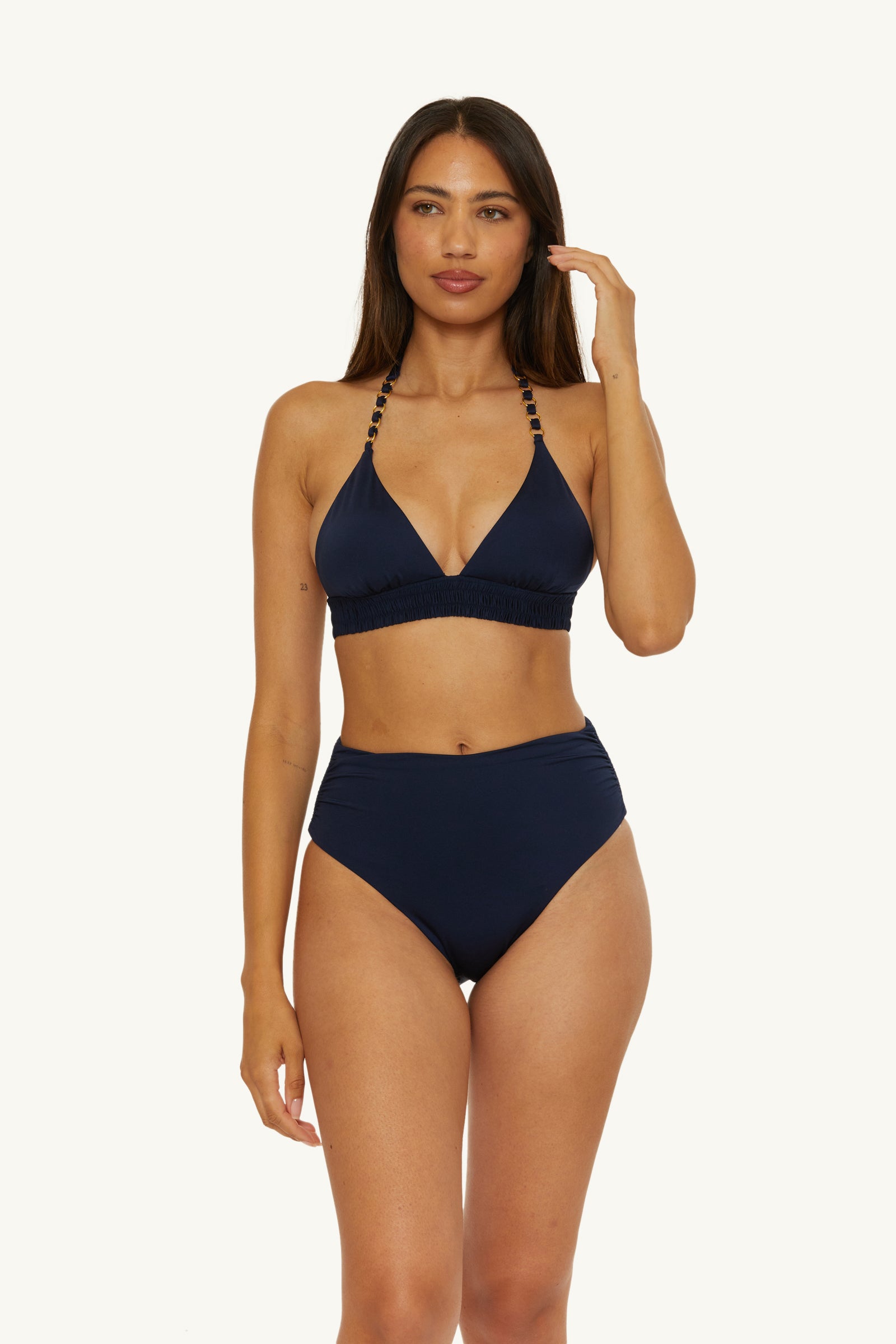 A woman stands against a plain light background, wearing the Shirred High-Waist Swim Bottom - Monaco and a navy blue bikini top with beaded straps, looking forward with one hand near her hair.