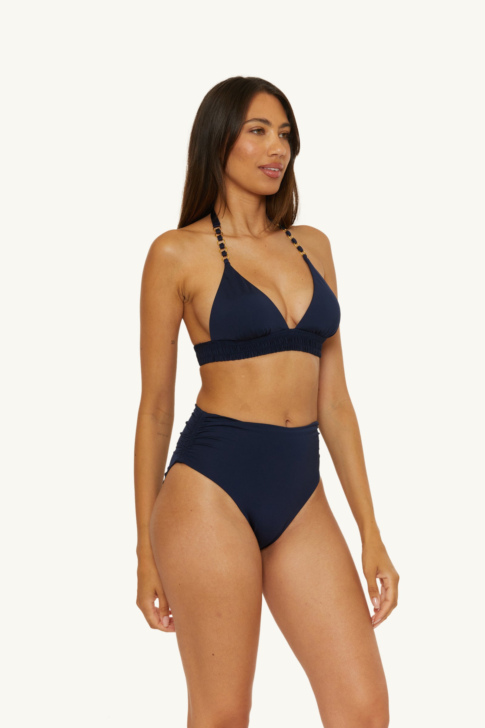 A woman with long brown hair models the Ring Halter Swim Top - Monaco in navy blue, featuring bead details and paired with high-waisted bottoms, against a plain background. She looks relaxed, gazing slightly to the side.