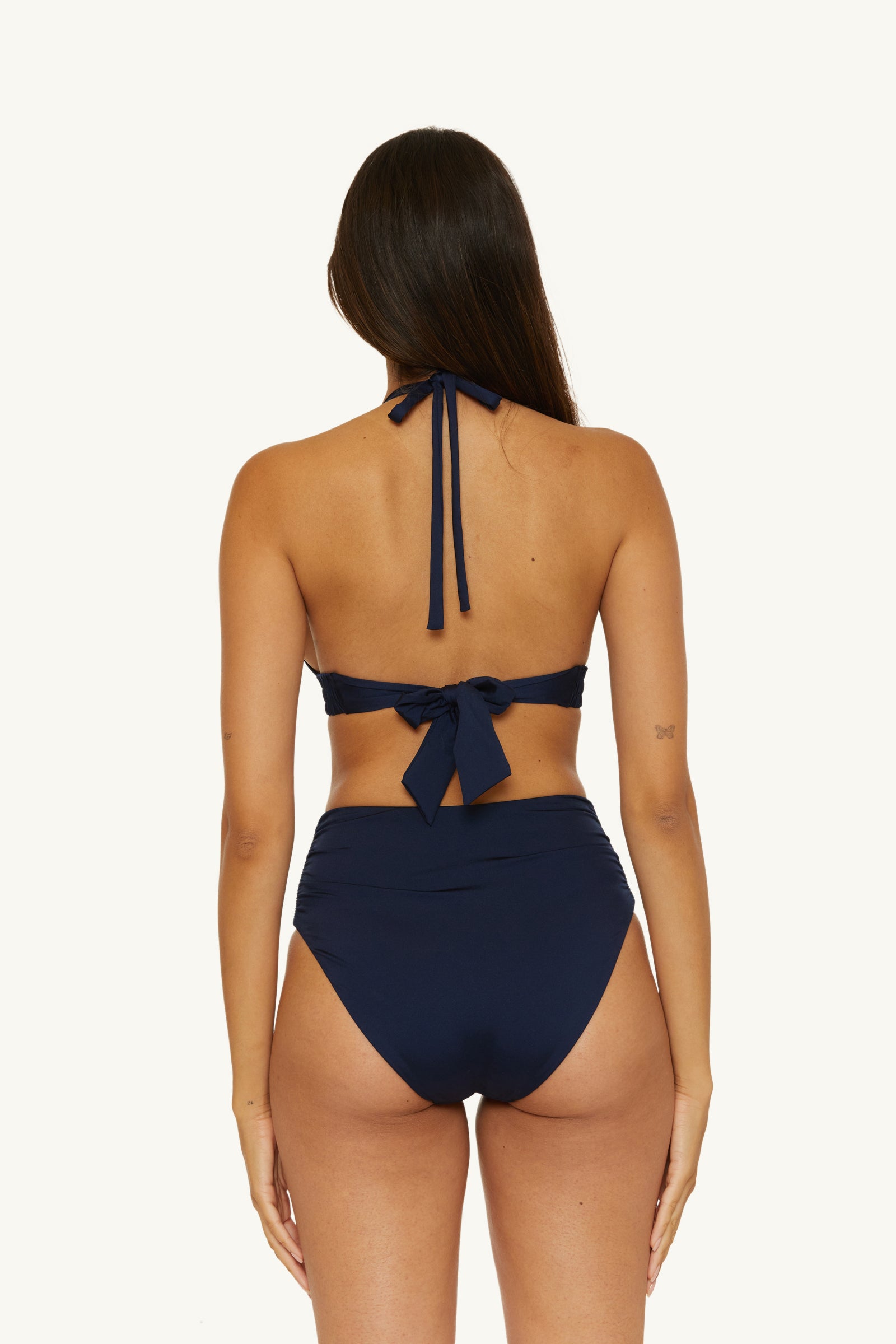 A woman with long brown hair stands facing away, wearing an ink-colored halter-neck bikini top and the Shirred High-Waist Swim Bottom - Monaco. The background is plain white.