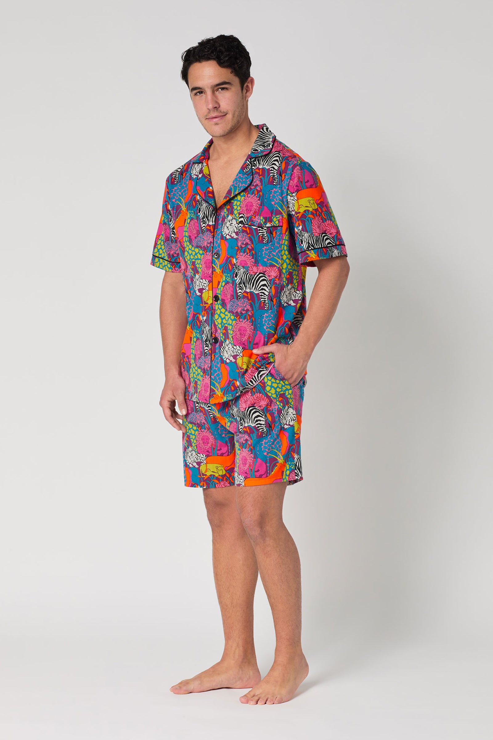 A man stands barefoot against a white background, wearing the Men's Boxer PJ Set - Jungle: a vibrant short-sleeve button-up shirt and matching shorts featuring bold multicolored animal and floral prints.