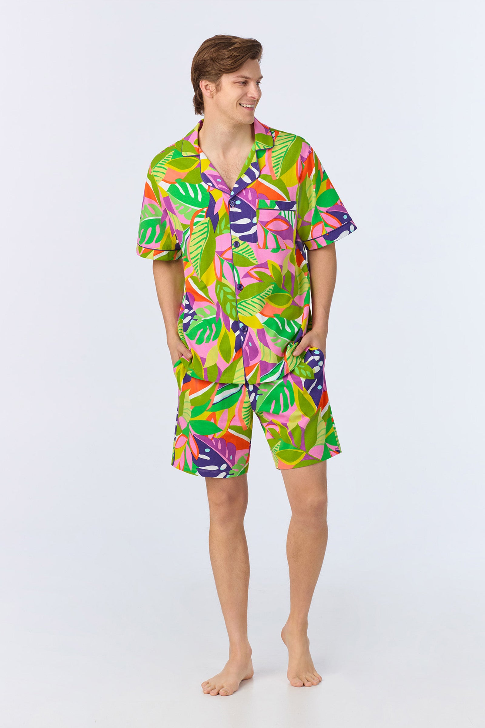 A smiling man stands barefoot, wearing the Mens Boxer PJ Set - Jungle Stretch Jersey in Multi, featuring a short-sleeve shirt and matching shorts with a bold tropical leaf pattern on a light background.