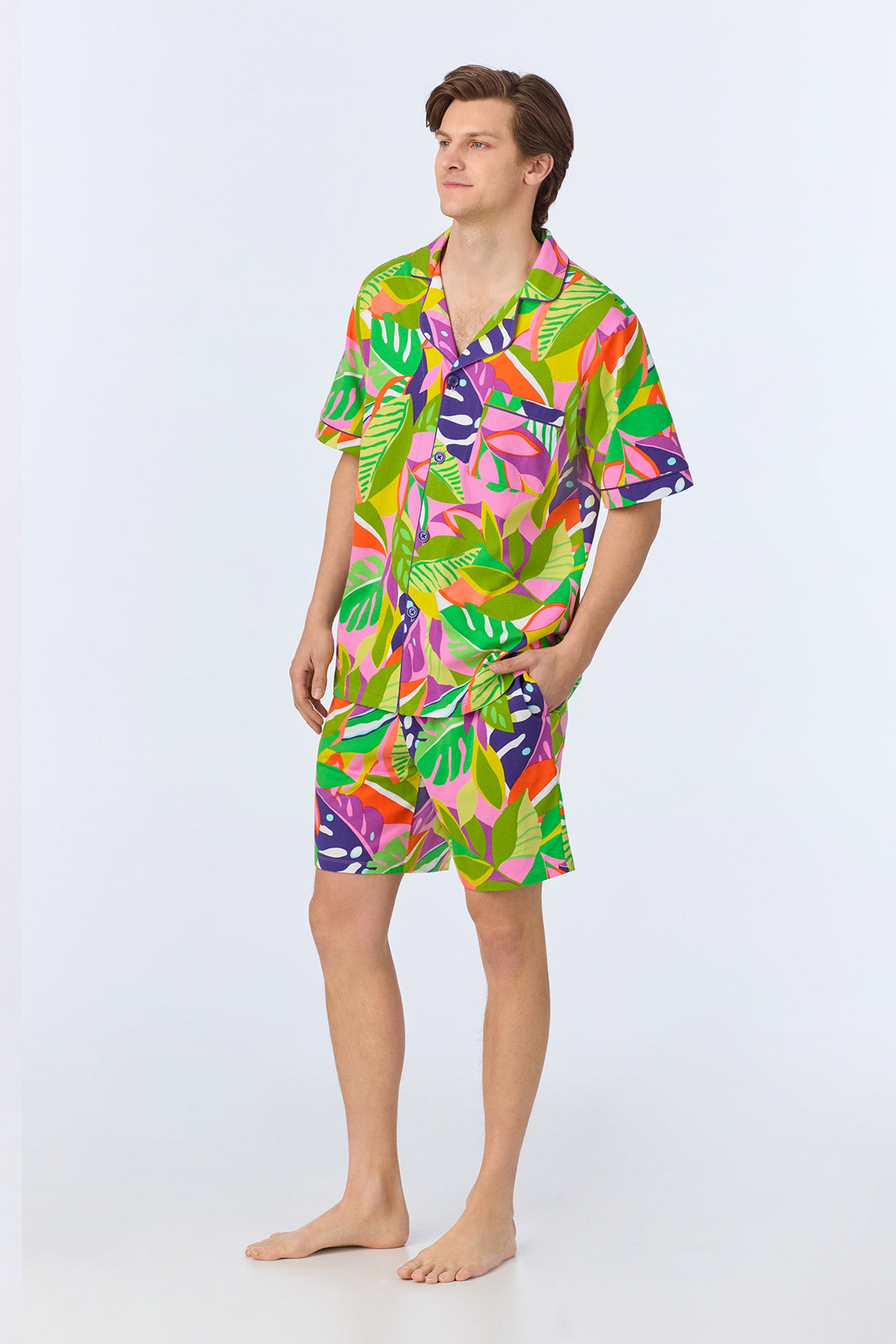 A person stands barefoot in the Mens Boxer PJ Set - Jungle Stretch Jersey, featuring a matching short-sleeve shirt and shorts with a vibrant, multicolored tropical leaf pattern against a plain light background.