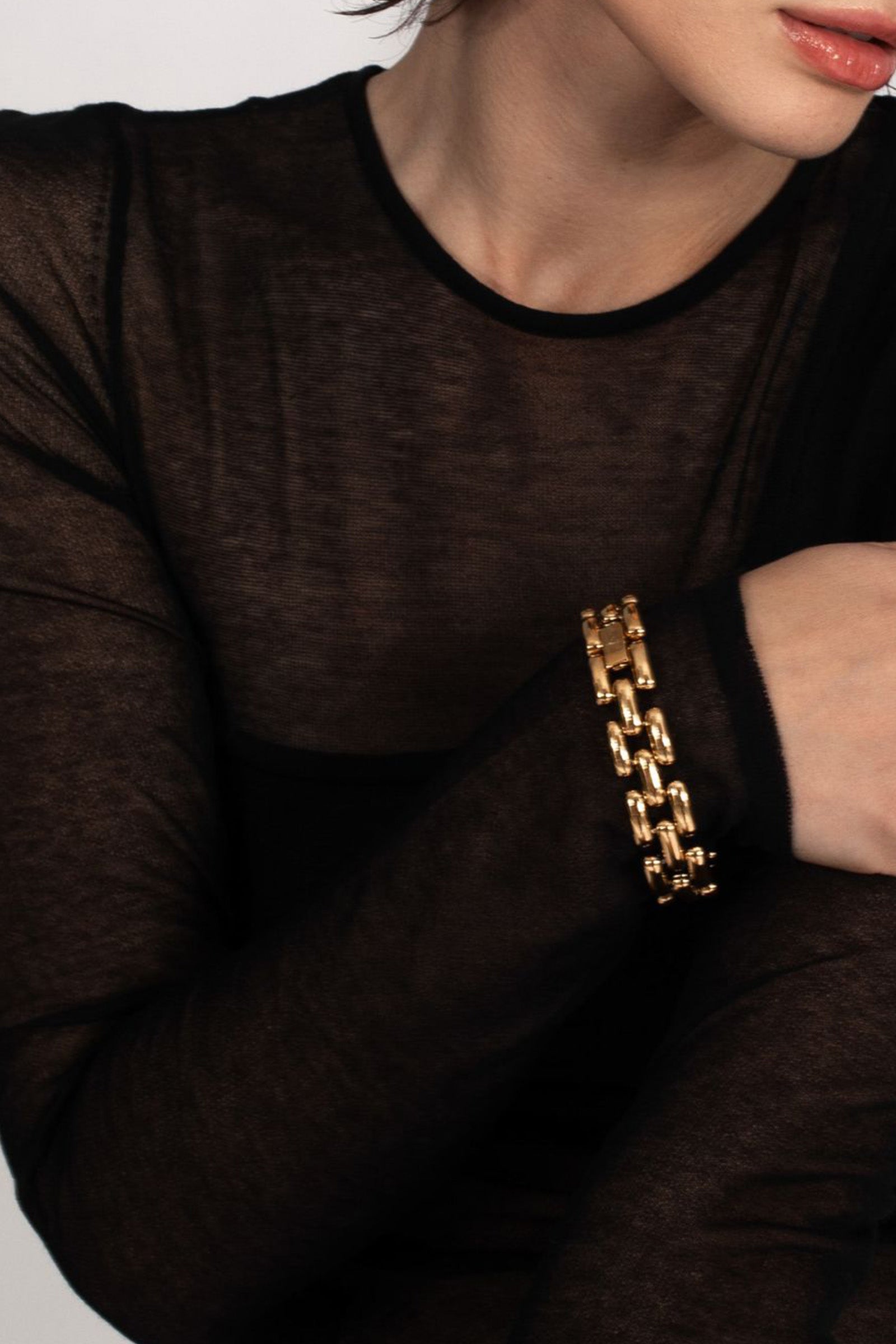 A person wears the Phoebe Bracelet, a chunky gold chain, on their wrist with a sheer black long-sleeve top. Their arm is bent and hand rests near their chest; their face is partially visible.