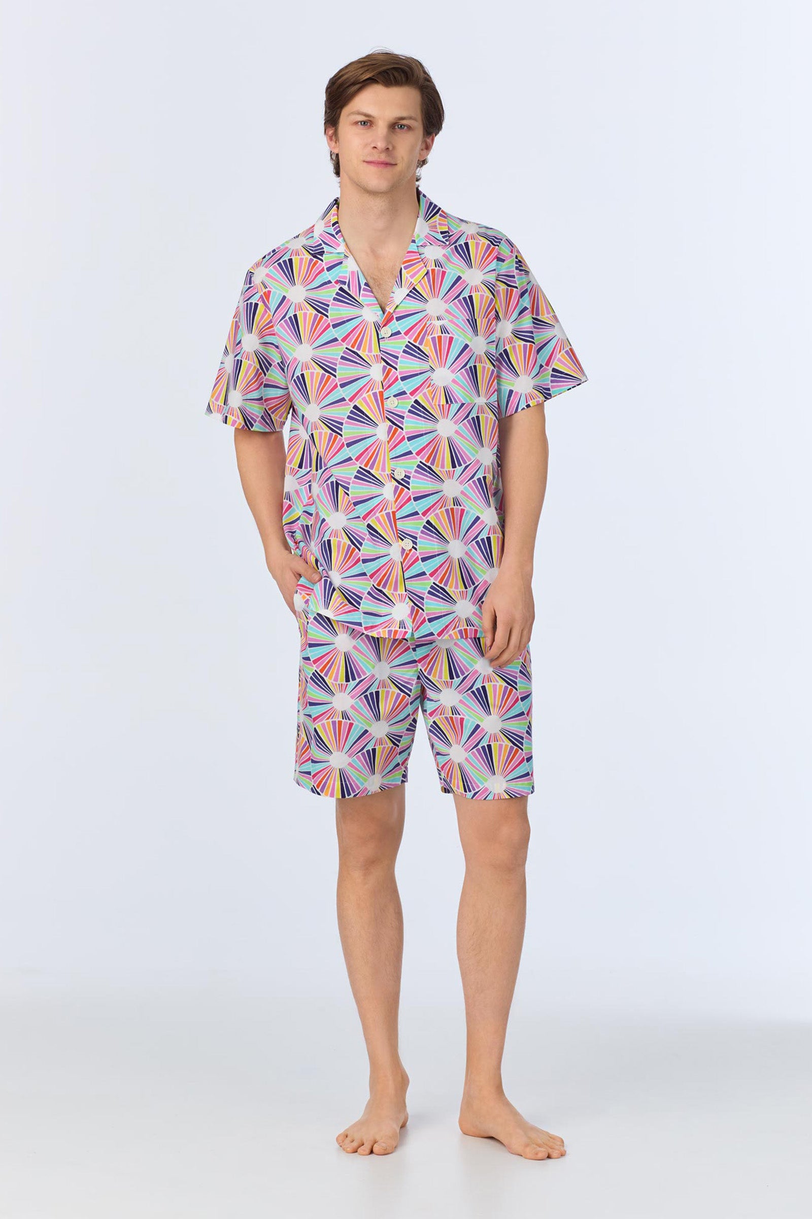A young man stands barefoot against a plain light background, wearing the Mens Boxer PJ Set - Fanfare Poplin in Multi, featuring a matching short-sleeved button-up shirt and shorts with a colorful geometric fan pattern.