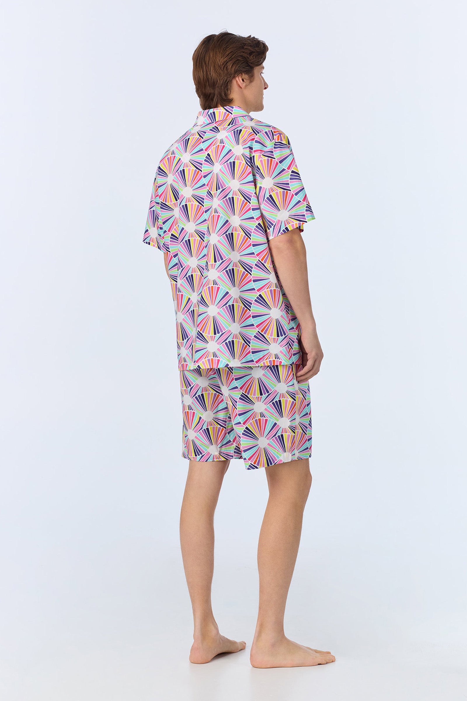 A barefoot man stands facing away, wearing the Mens Boxer PJ Set - Fanfare Poplin, which features a colorful geometric fan pattern on a light background in a matching short-sleeve shirt and shorts.