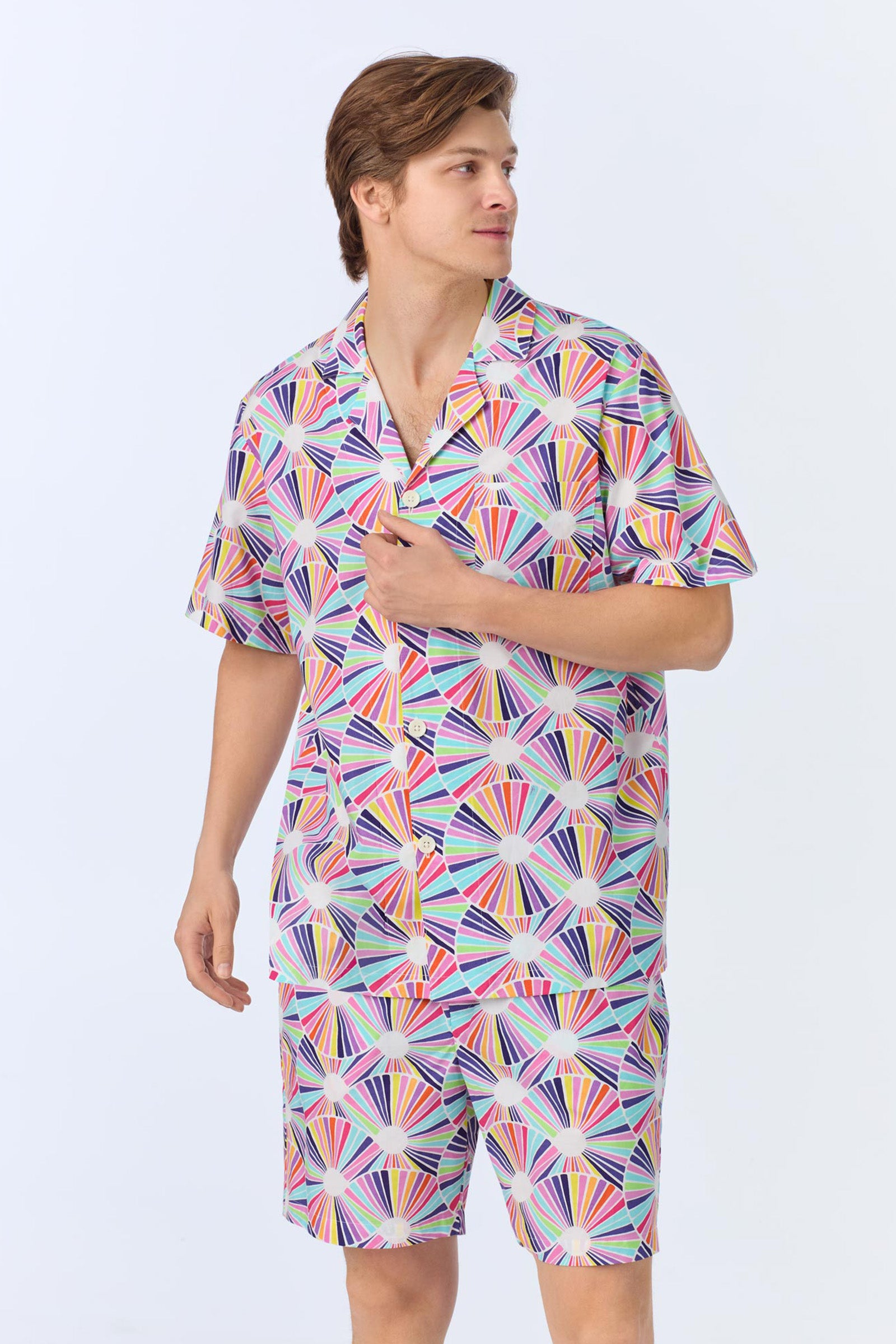 A person wears the Mens Boxer PJ Set - Fanfare Poplin, a matching button-up shirt and shorts with a colorful geometric fan print in pink, purple, yellow, blue, and white against a plain light background.