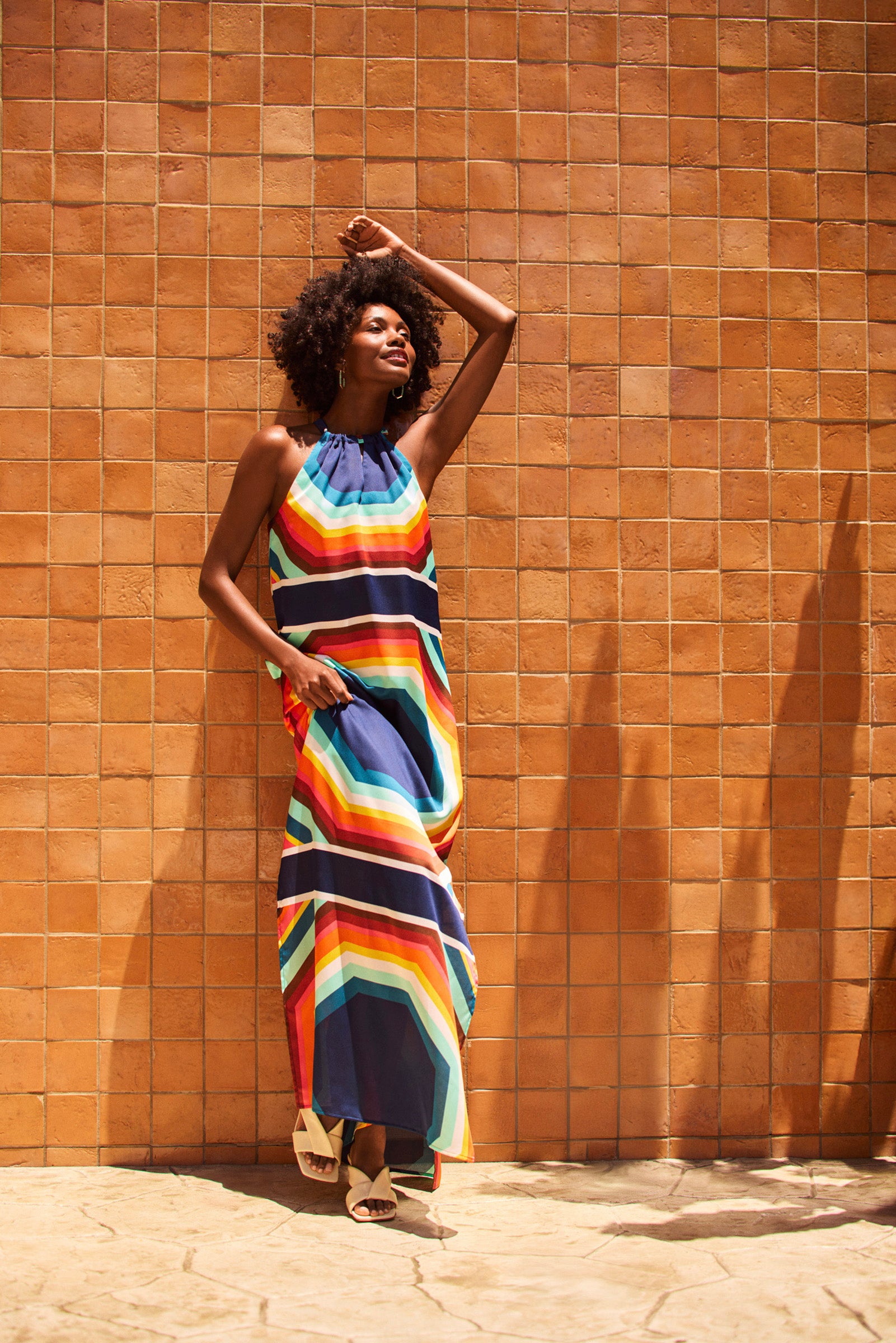 A woman wearing the Emilla Sleeveless Maxi Dress leans against a textured orange tile wall, smiling with one arm raised. She stands in bright sunlight, wearing white sandals and casting soft shadows.