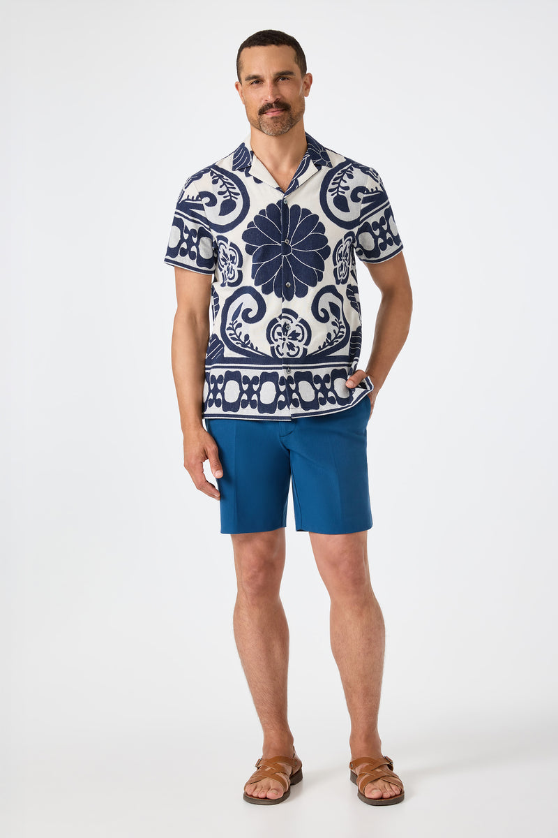 A man with a mustache wears the Felix Camp Shirt - Fuyuko, blue shorts, and brown sandals while standing against a plain white background.
