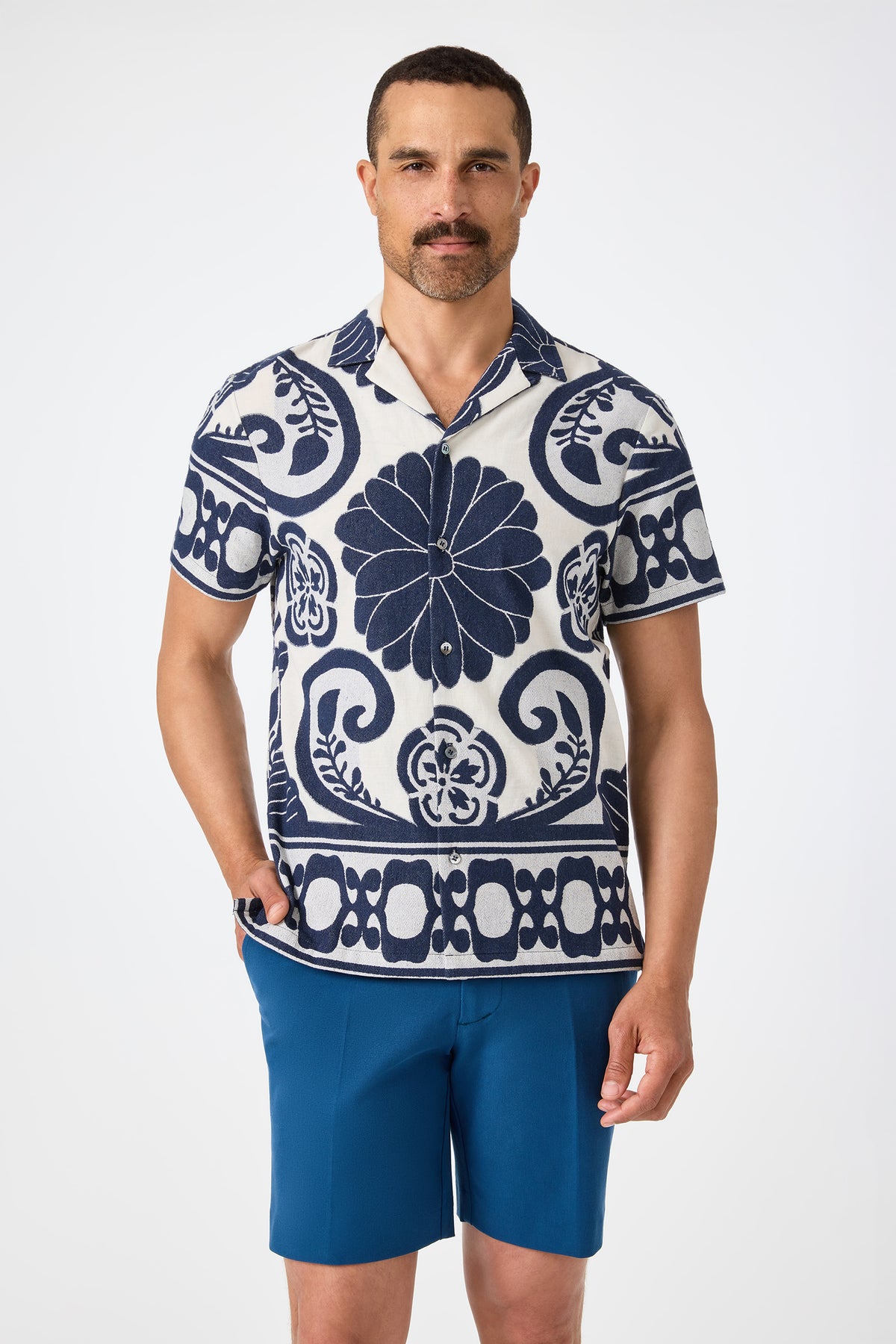 A man with short dark hair and a mustache stands against a plain background wearing the Felix Camp Shirt in Fuyuko Fil Coupe, featuring bold navy blue floral and geometric patterns, paired with solid blue shorts and one hand in his pocket.