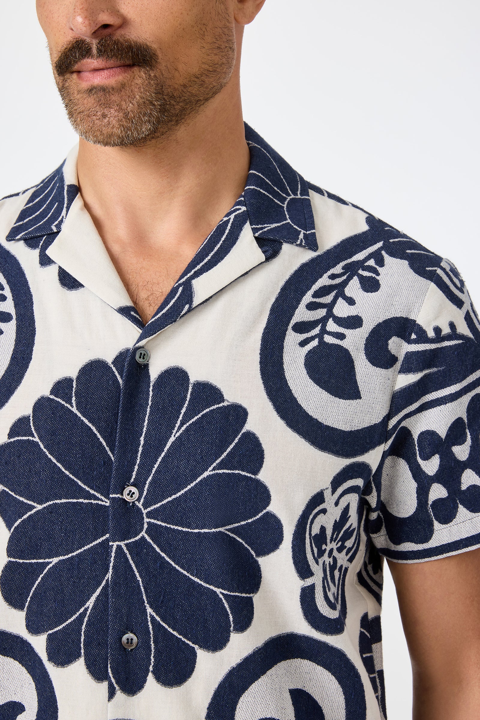 A man with a trimmed beard wears the Felix Camp Shirt - Fuyuko, featuring bold navy floral and abstract prints on light beige. The photo shows his upper torso and part of his face.