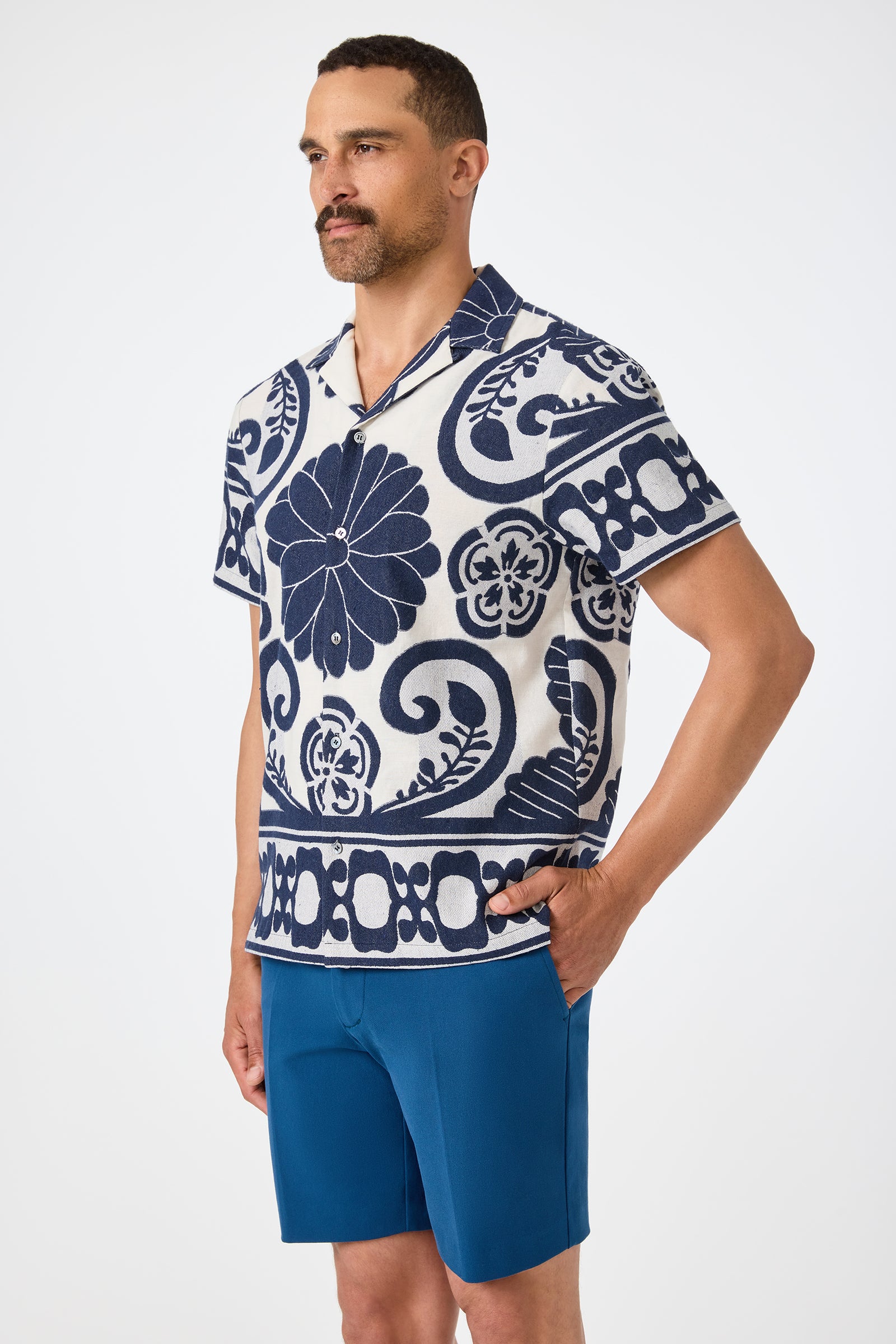 A man stands with his hands in his pockets, wearing the Felix Camp Shirt - Fuyuko, a short-sleeve collared shirt with a bold blue and white floral pattern, and blue shorts, against a plain white background.