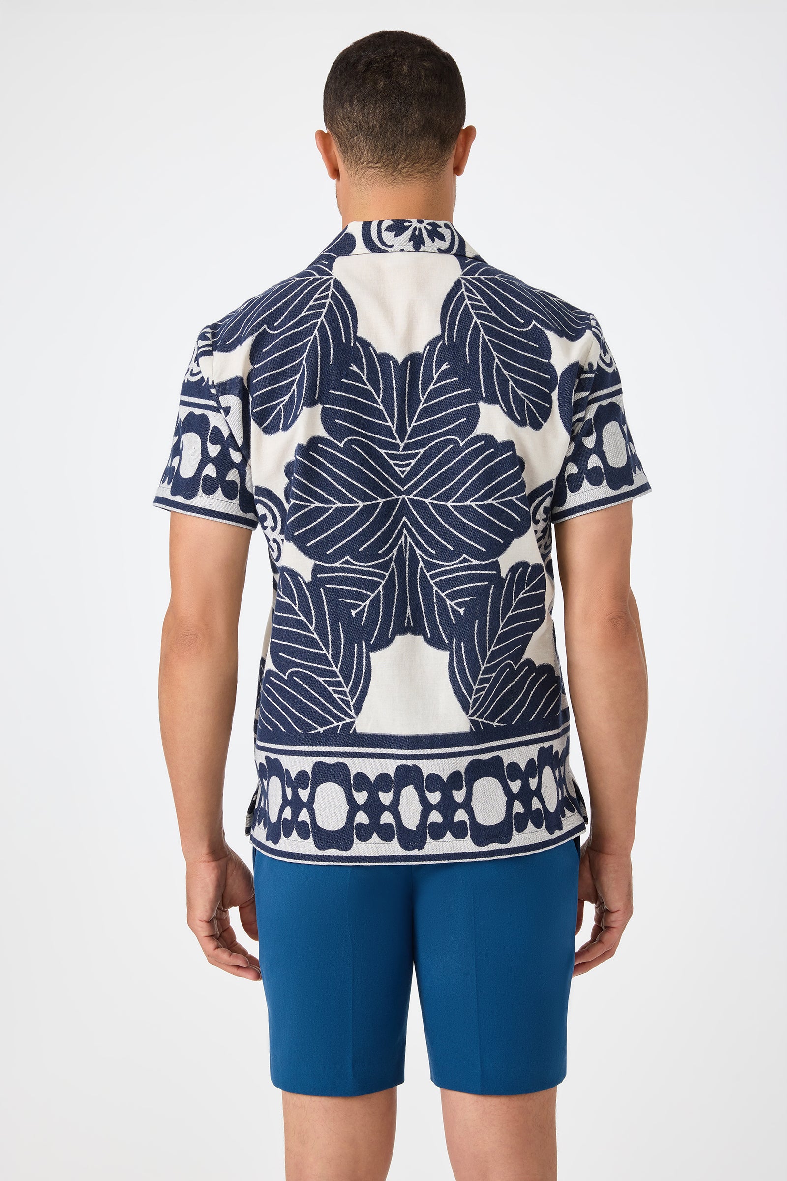 A man faces away from the camera wearing the Felix Camp Shirt - Fuyuko, featuring bold navy blue leaf and geometric patterns on white, paired with solid blue shorts against a plain white background.
