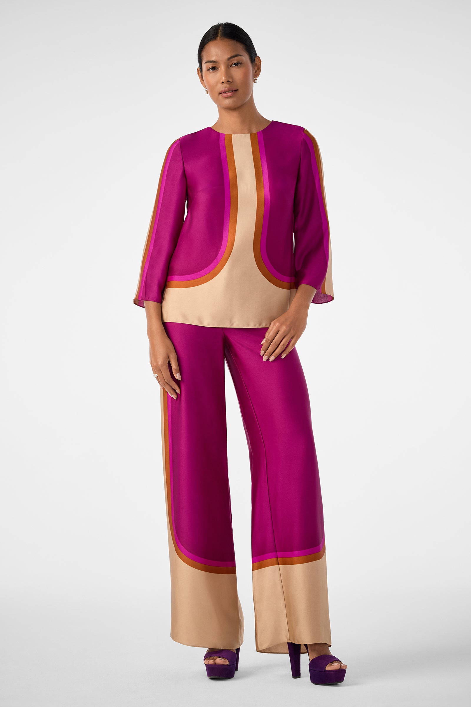 A woman wears the Verity 2 Pant, a wide-leg style with a bold geometric pattern in magenta, tan, and red. She pairs it with a matching long-sleeve top and purple platform heels against a plain background.