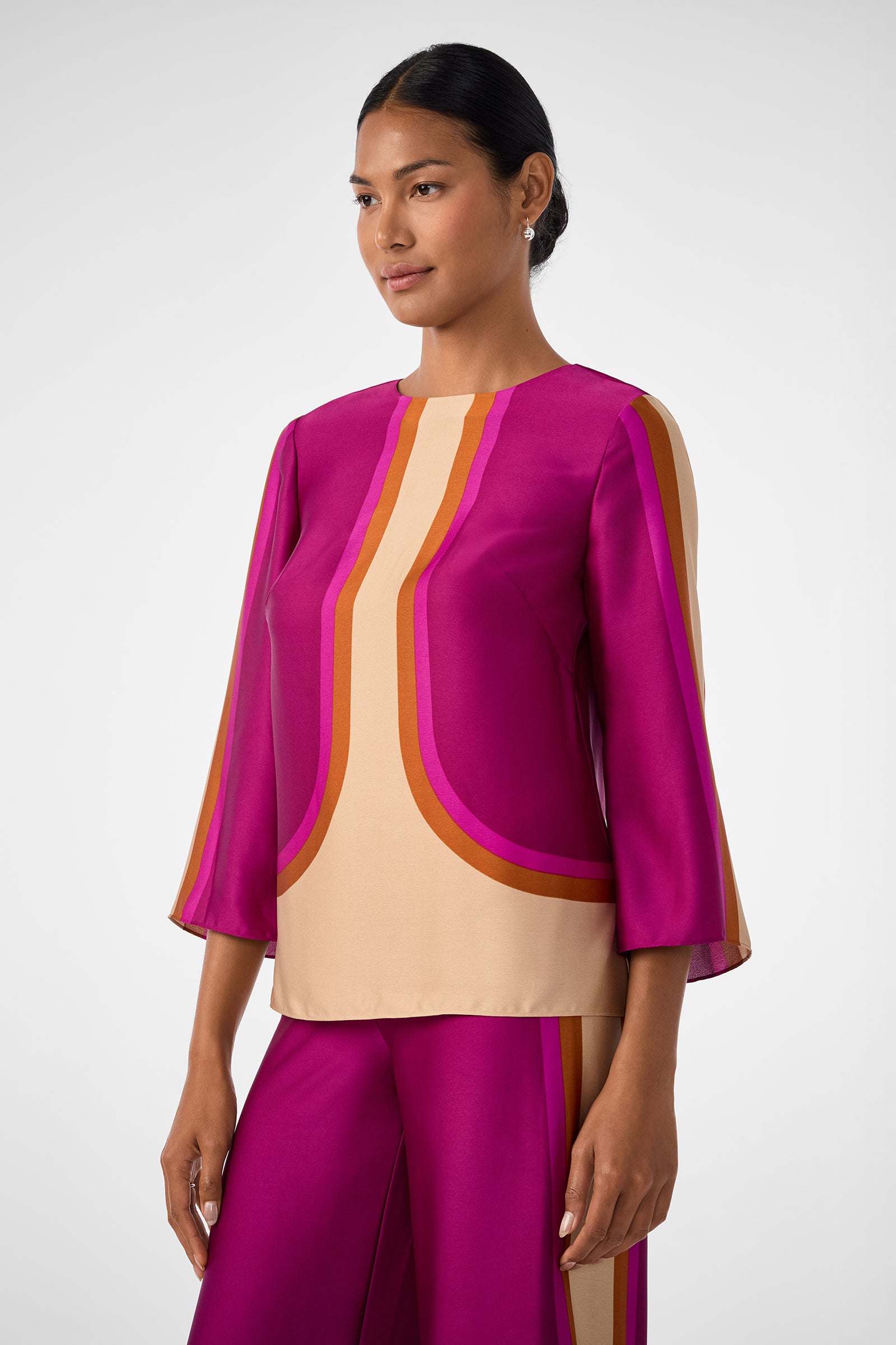 A woman wears the Sprig 3 Top, a matching magenta, beige, and orange patterned piece with three-quarter sleeves and wide-leg pants. She stands against a plain white background, looking slightly to the side with a neutral expression.
