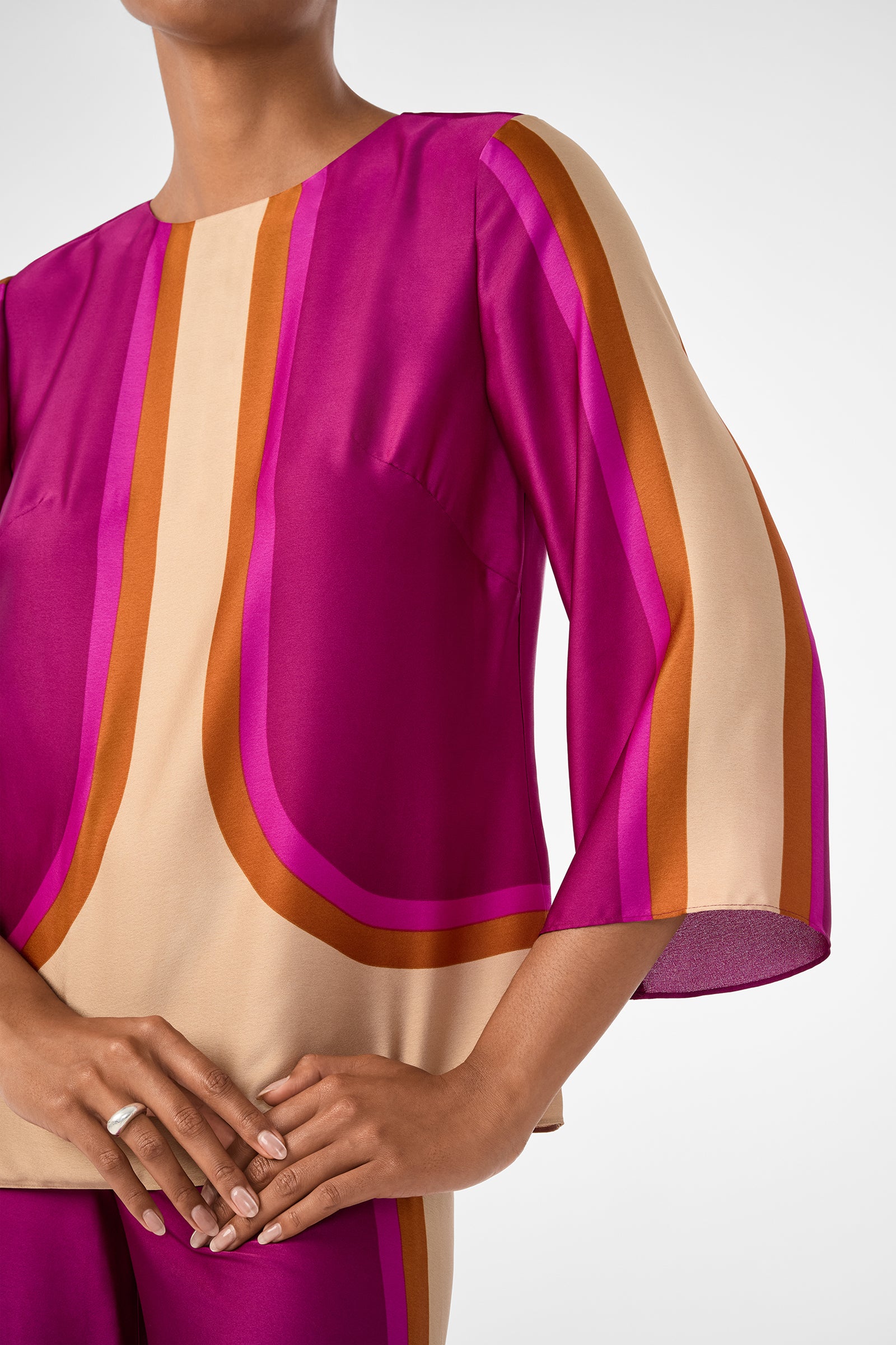 Wearing the Sprig 3 Top—a vibrant blouse with wide sleeves and bold magenta, orange, and beige geometric stripes—the persons hand rests on their hip, upper body visible against a plain background.