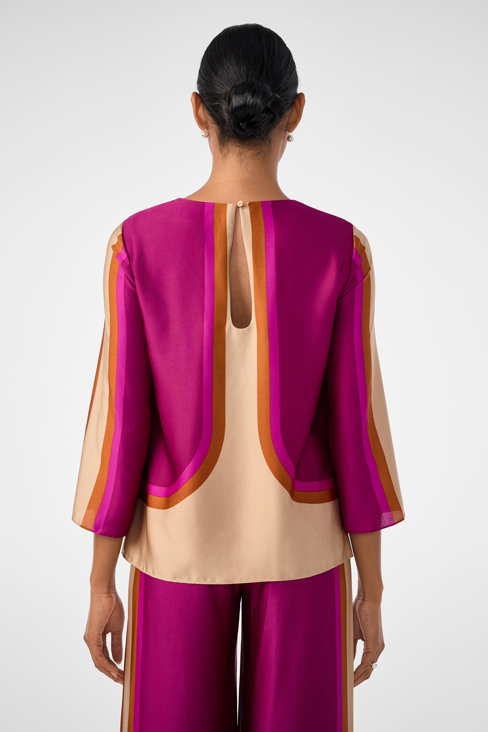 A person with dark, neatly tied hair is seen from behind wearing the Sprig 3 Top—a vibrant, long-sleeved blouse in purple, beige, and orange with bold geometric stripes and a keyhole opening at the back.